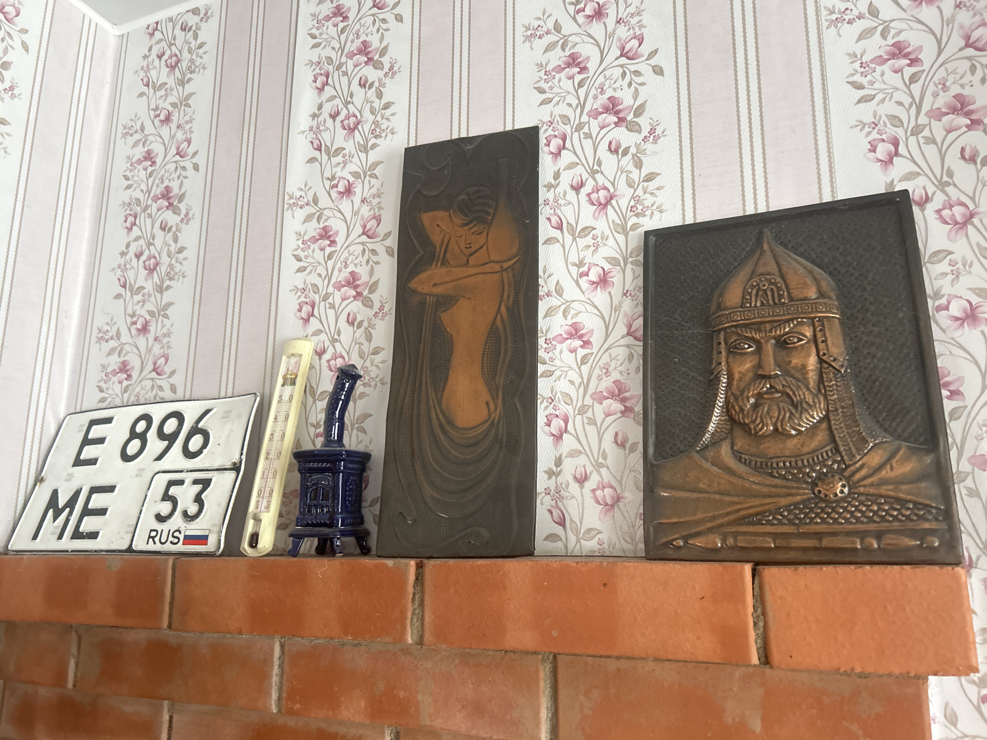 The image shows a shelf on a brick fireplace. On the shelf, from left to right, are a license plate, a thermometer, a small blue ceramic stove, a dark rectangular relief of a woman, and a dark rectangular relief of a man in armor. The license plate has the characters "E 896" and "ME 53" along with "RUS" and the Russian flag. The thermometer is yellow and has markings for temperature. The blue ceramic stove is detailed and appears to be a decorative item. The relief of the woman shows her in a stylized pose, and the relief of the man is a bust of a bearded figure wearing a helmet and armor. The background is wallpaper with a floral pattern and vertical stripes. The fireplace is made of orange bricks.