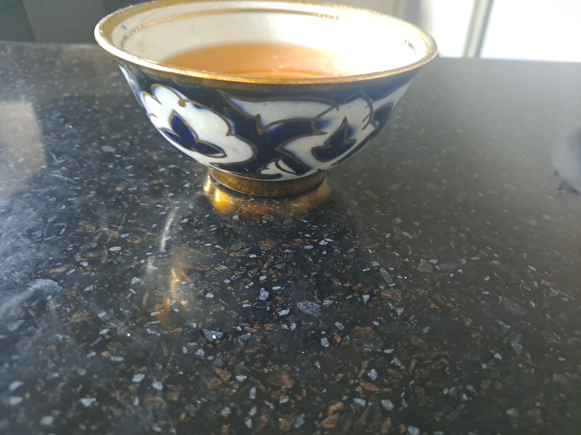 The image shows a close-up of a decorative porcelain teacup filled with a golden-amber liquid, likely tea. The teacup has a white interior and is adorned with intricate blue and white floral patterns with gold trim around the rim and on its small foot. It rests on a dark, speckled countertop that appears to be granite or a similar composite material, with flecks of gray, black, and brown. The lighting suggests an indoor setting, possibly near a window, as indicated by the diffuse light and slight reflections on the countertop. The background is blurred, but hints of a bright, possibly outdoor or windowed area are visible. There are no people or specific urban landmarks identifiable that would indicate a location within Namangan City, Uzbekistan.