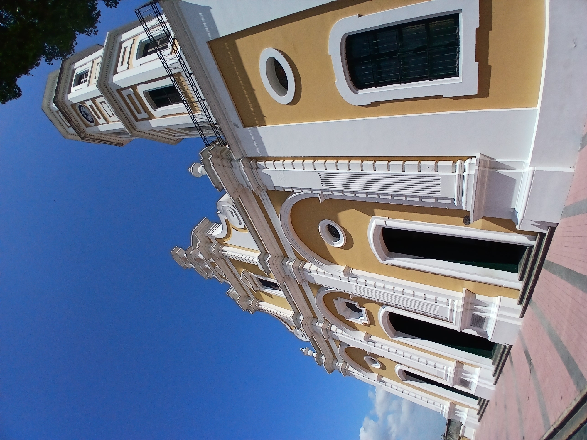 The image shows the Cathedral of Ciudad Bolivar in Venezuela. The cathedral has a yellowish facade with white accents and a bell tower on the left side. The sky is clear and blue. The image is taken from a low angle, looking up at the building. The architecture is colonial style. The ground is paved with red and gray bricks.