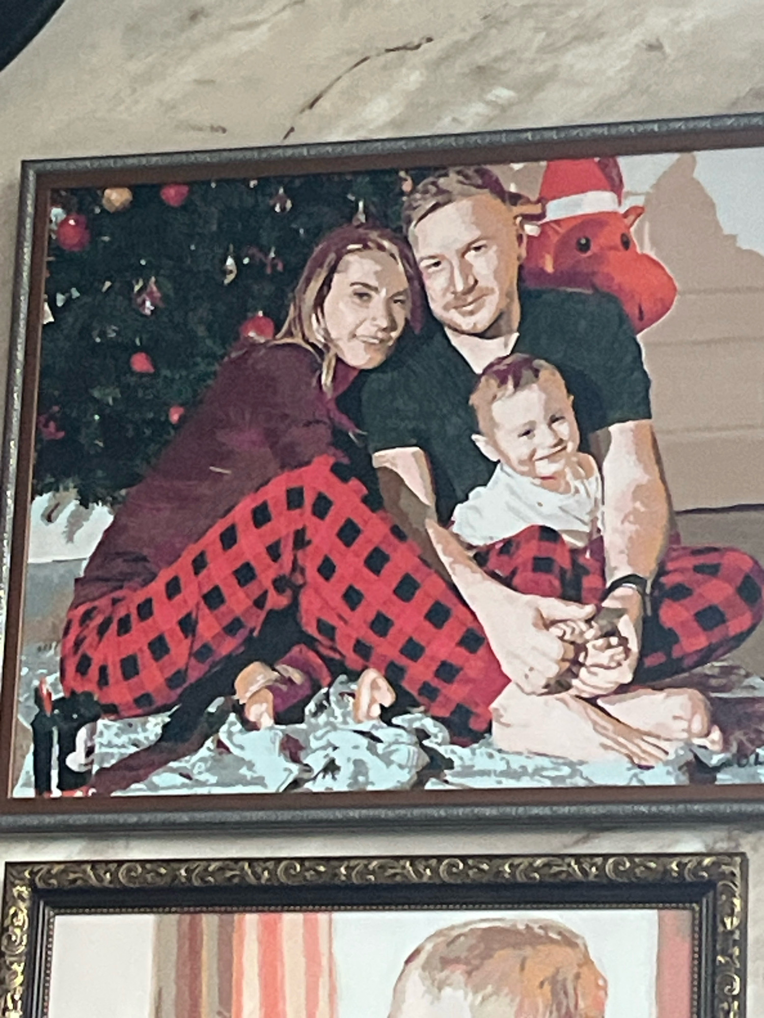 The image is a painting of a family of three, a man, a woman, and a young child, posed in front of a Christmas tree. They are all wearing red and black plaid pajamas. The man is on the right, holding the child on his lap. The woman is on the left, leaning on the man. The child is smiling and looking at the camera. The Christmas tree is decorated with red ornaments and lights. There is also a red stuffed animal with a Santa hat behind the man. The painting is framed in a dark wood frame. There is another framed painting below the first one. This image is not located in the city of Vladimirovka, Russia.
