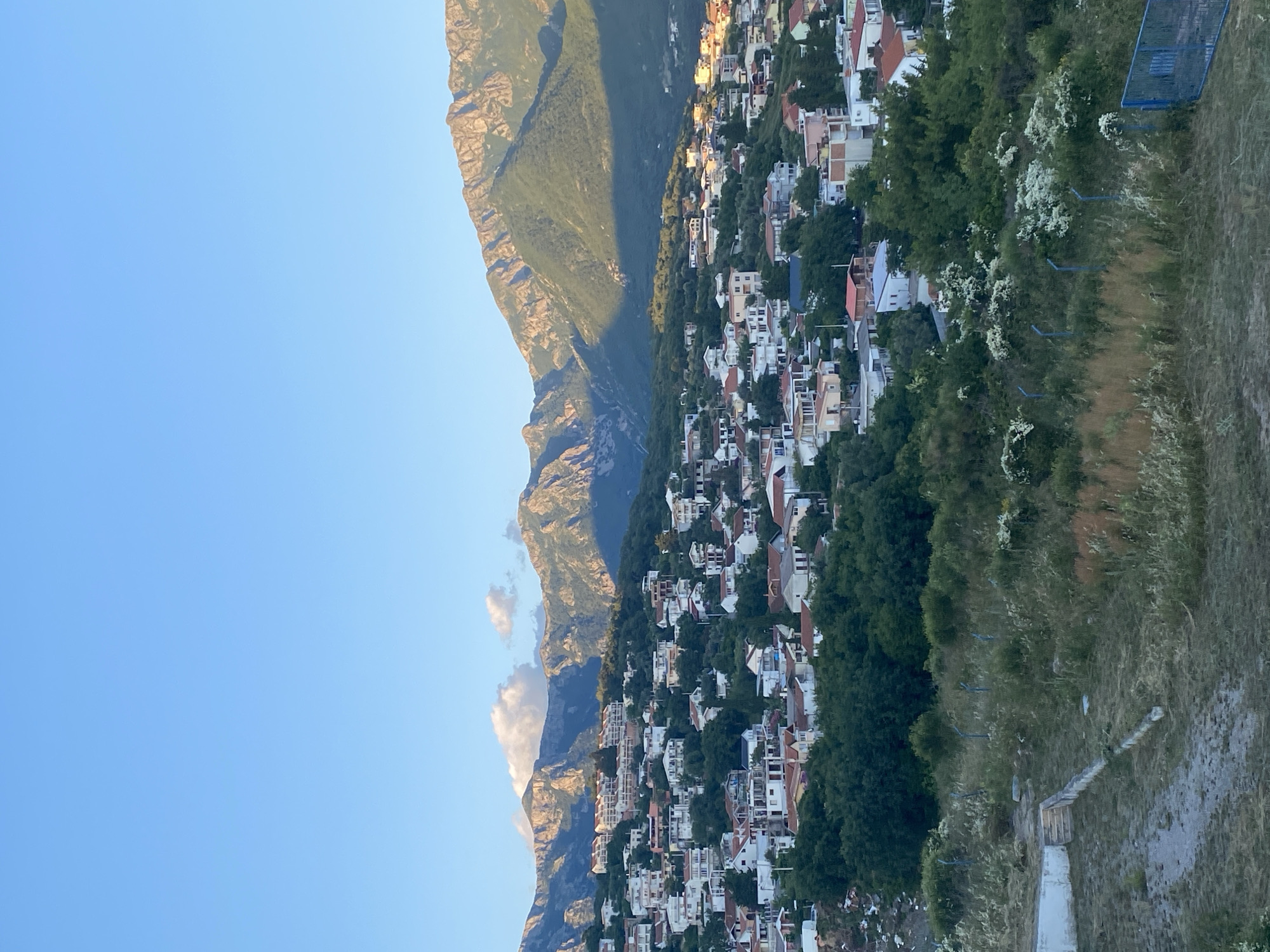 The image shows a panoramic view of a hillside town in Šušanj, Montenegro, under a clear blue sky. The foreground features a grassy area with some dry vegetation and a low concrete structure with steps. A thin wire fence runs horizontally across the middle ground, separating this area from the densely packed houses of the town.
The town itself is characterized by numerous buildings with red-tiled roofs, interspersed with lush green trees. The houses are of varying sizes and architectural styles, suggesting a mix of residential properties. The town extends up the gentle slope of the hill, gradually merging with the surrounding vegetation.
In the background, majestic mountains rise dramatically, their rugged peaks illuminated by the warm light of what appears to be late afternoon or early evening. The mountains are partially covered in green vegetation, with some rocky outcrops visible. Shadows are cast across the mountain slopes, indicating the direction of the sun. A few wispy clouds are present in the sky above the mountains. There are no people or animals visible in the scene. The overall impression is one of a peaceful, sun-drenched settlement nestled between the sea (implied but not visible) and the mountains.