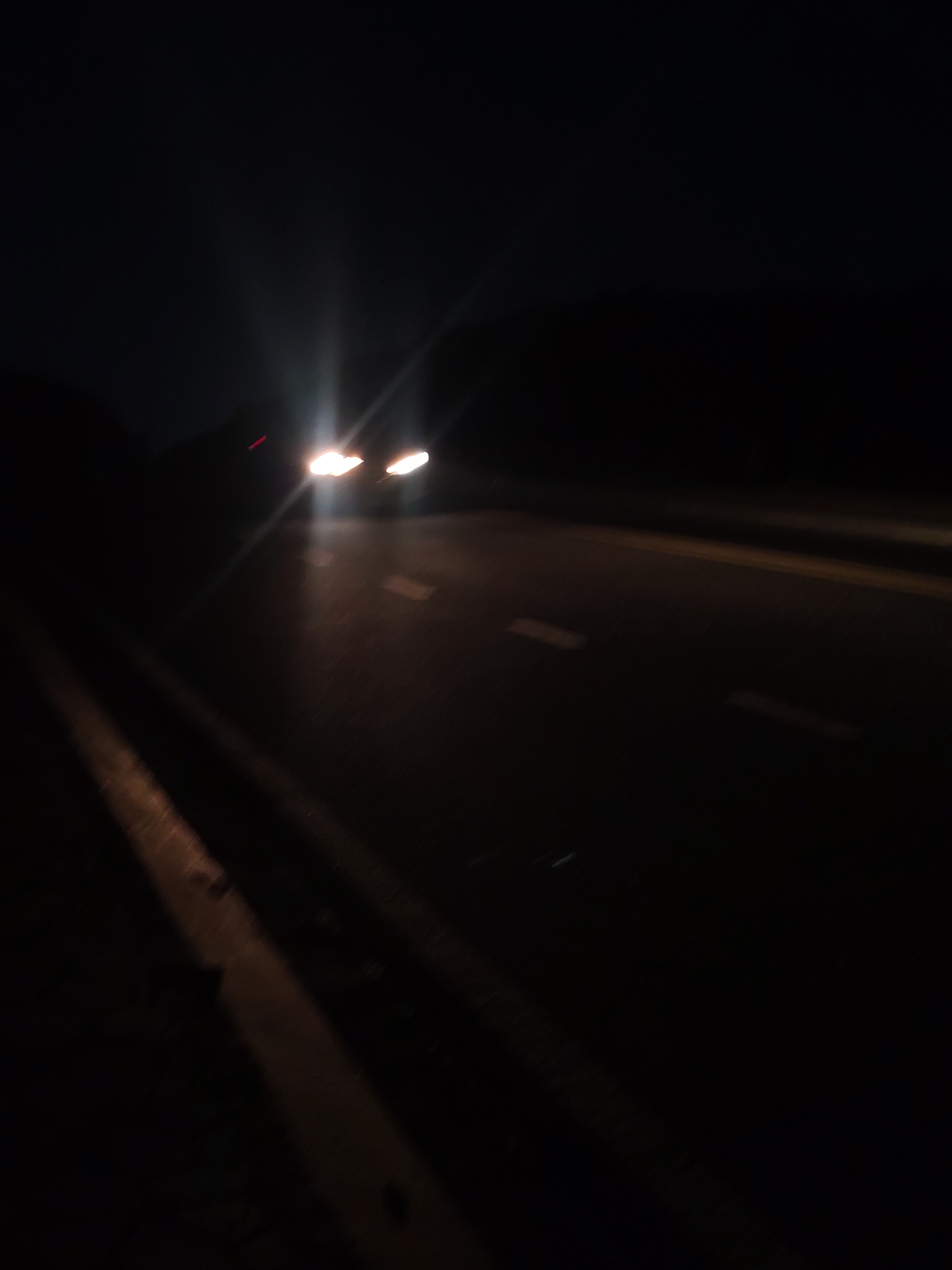 A long exposure photograph captures a dark, blurry scene of a road at night, with the headlights of an approaching vehicle creating streaks of light across the asphalt. The vehicle appears to be moving towards the viewer, indicated by the bright, elongated headlights. White dashed lines marking the lanes are visible on the road surface, appearing smudged due to motion. The left side of the image shows a dark embankment or roadside, with a faint, illuminated stripe running parallel to the road, possibly a reflective barrier or edge marking. The overall impression is one of speed and low visibility, common on highways or less populated roads at night. The image does not contain specific landmarks or features that would identify it as being in Da Lat, Vietnam, beyond the general context of a road at night.