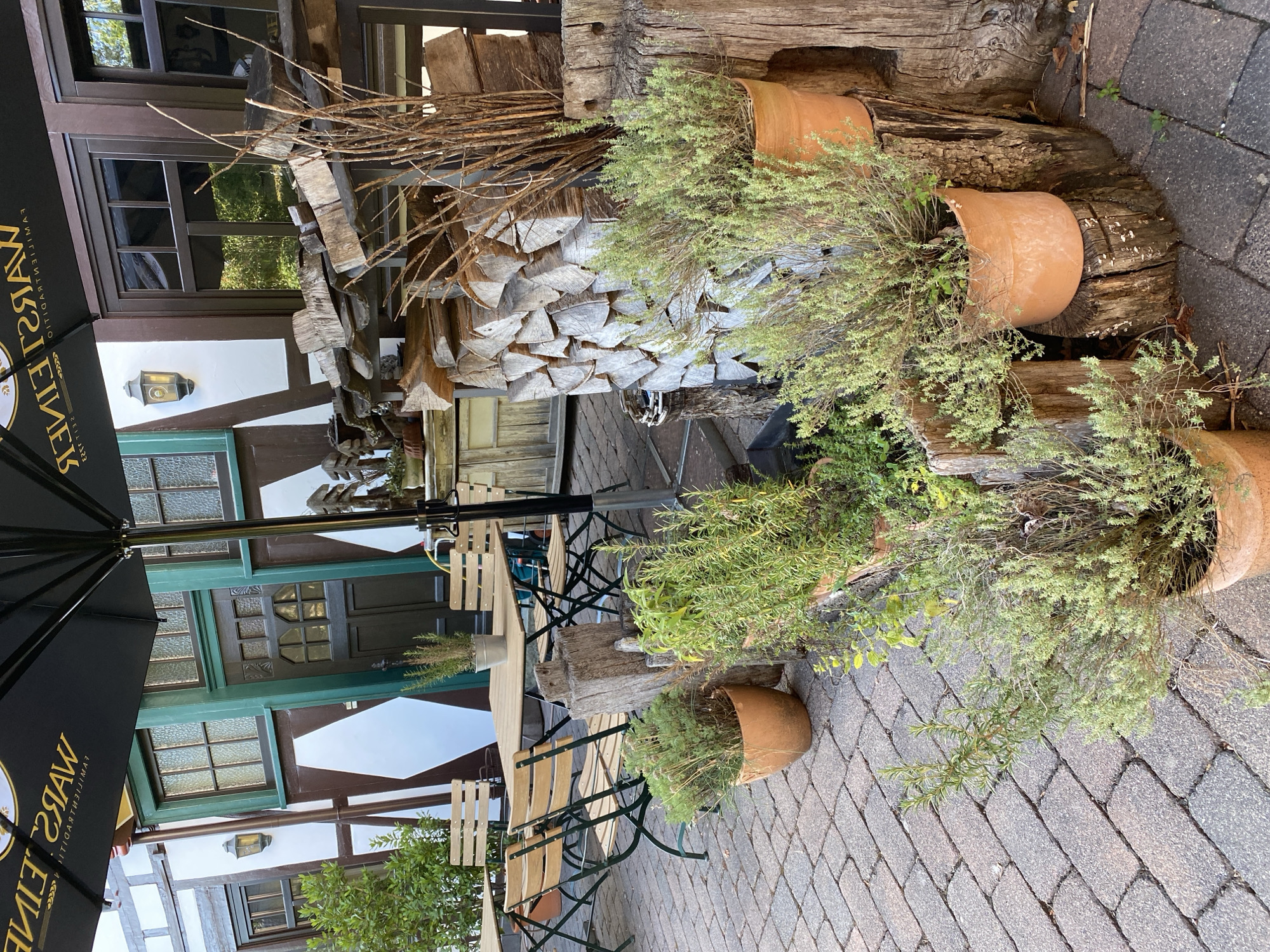 The image shows an outdoor seating area of a restaurant or cafe in Seelbach bei Hamm (Sieg), Germany. A black umbrella with text on it is partially visible in the upper portion of the frame. In the background, a building with traditional German half-timbered architecture is visible, featuring dark wood beams, white infill, and green-trimmed windows and doors.

In the foreground and mid-ground, several wooden tables and chairs are set up on a paved patio made of brick-like stones. There are several potted plants, including rosemary and other herbs, scattered throughout the seating area, some placed on wooden stumps. A stack of firewood is also visible, adding to the rustic ambiance. The scene is set during daylight, with sunlight casting shadows on the ground. No people are present in the image.