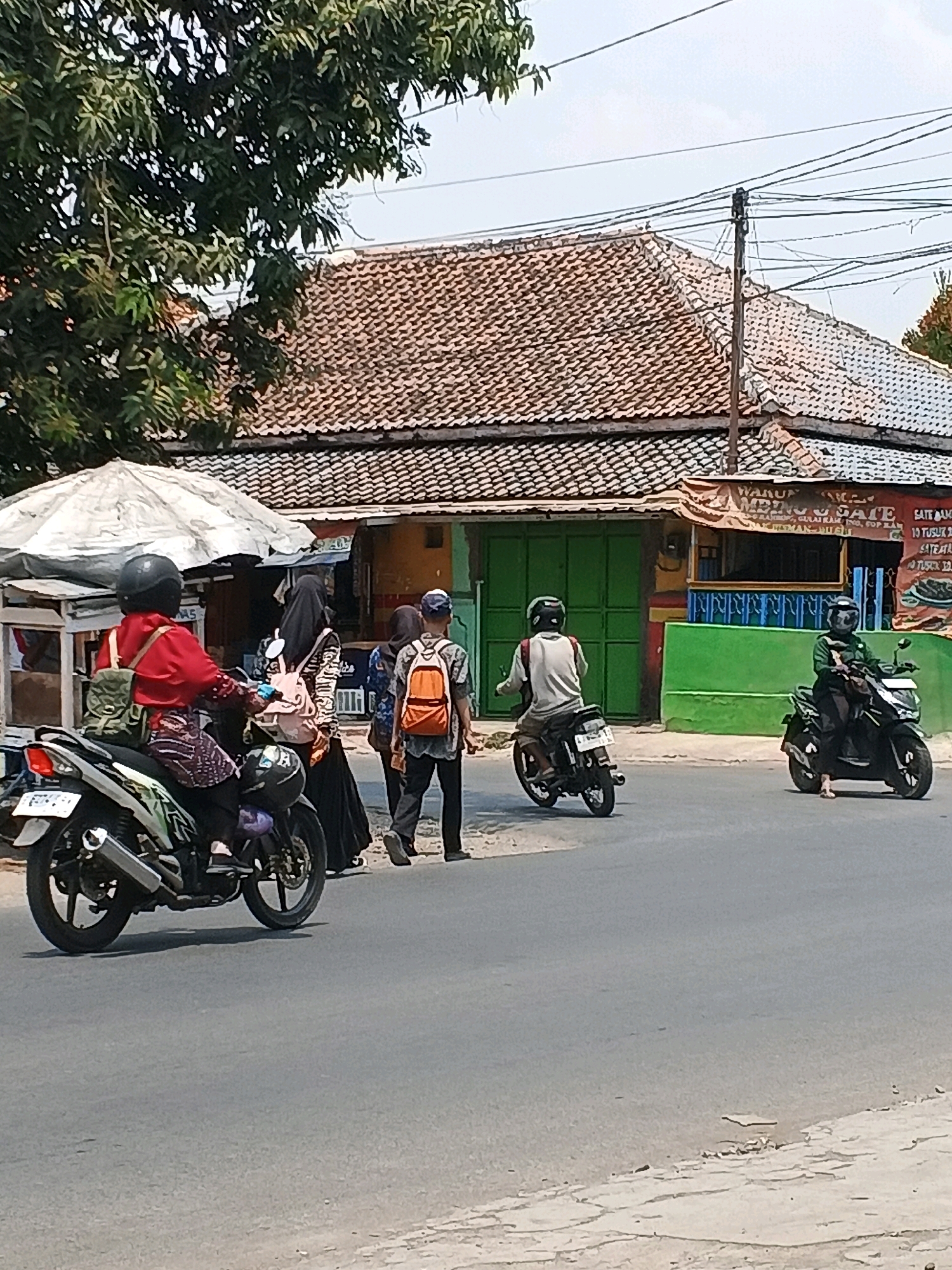 The image depicts a street scene in Taman, Indonesia, likely during daytime with overcast skies. In the foreground, a paved road is visible with a rougher texture on the right edge, suggesting a shoulder. Several individuals are present, primarily on or near motorcycles. On the left, a person wearing a red shirt, helmet, and patterned leggings rides a white and green motorcycle. A green backpack is attached to the back of the motorcycle. To their right, two individuals wearing black headscarves and loose clothing are walking, one carrying a pink backpack. Further to the right, a person in a grey shirt and black pants with an orange backpack is walking away from the viewer. On the road, two more motorcycles are visible. One, a black motorcycle on the right, is ridden by a person wearing a green jacket and helmet. Another black motorcycle is further left, moving away from the viewer, ridden by a person in a light-colored shirt and helmet. In the background, a building with a brown tiled roof and green doors is situated. A light-colored awning extends from a structure to the left of the building. Signs with text are visible on the building, suggesting commercial activity. Power lines are strung across the upper portion of the image, supported by a pole. Trees with green foliage are present on the left side of the frame.