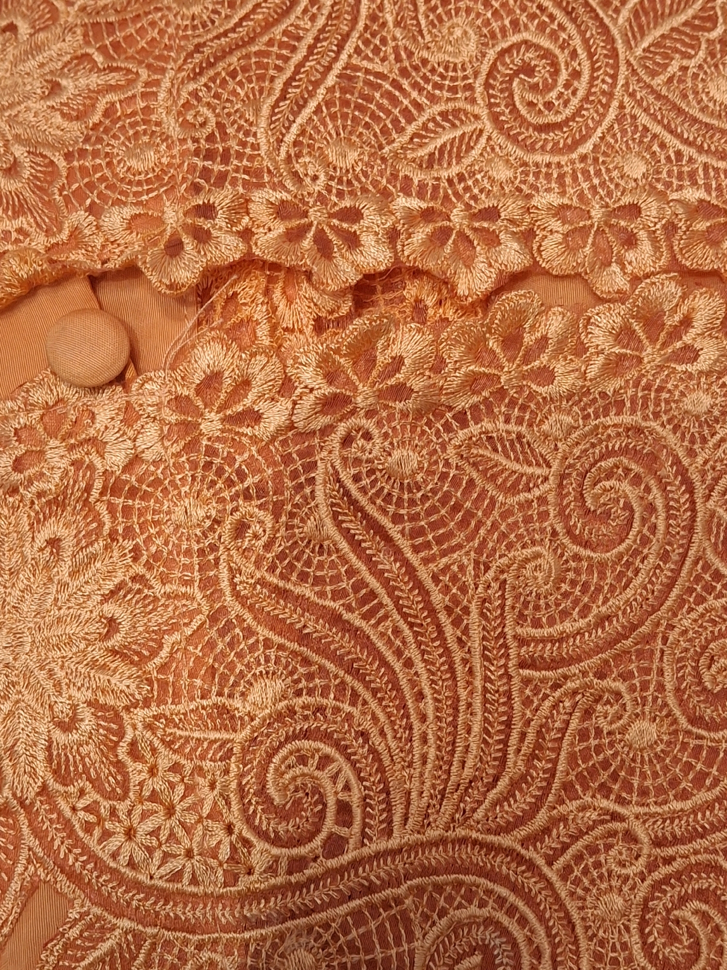 The image shows a close-up of an orange lace fabric with intricate embroidery. The pattern features swirling, floral, and geometric motifs. A small, round, orange button is visible in the upper left quadrant, partially obscured by the lace. The fabric appears to be a garment or textile. There is no discernible information within the image to determine a location in Lumajang, Indonesia.