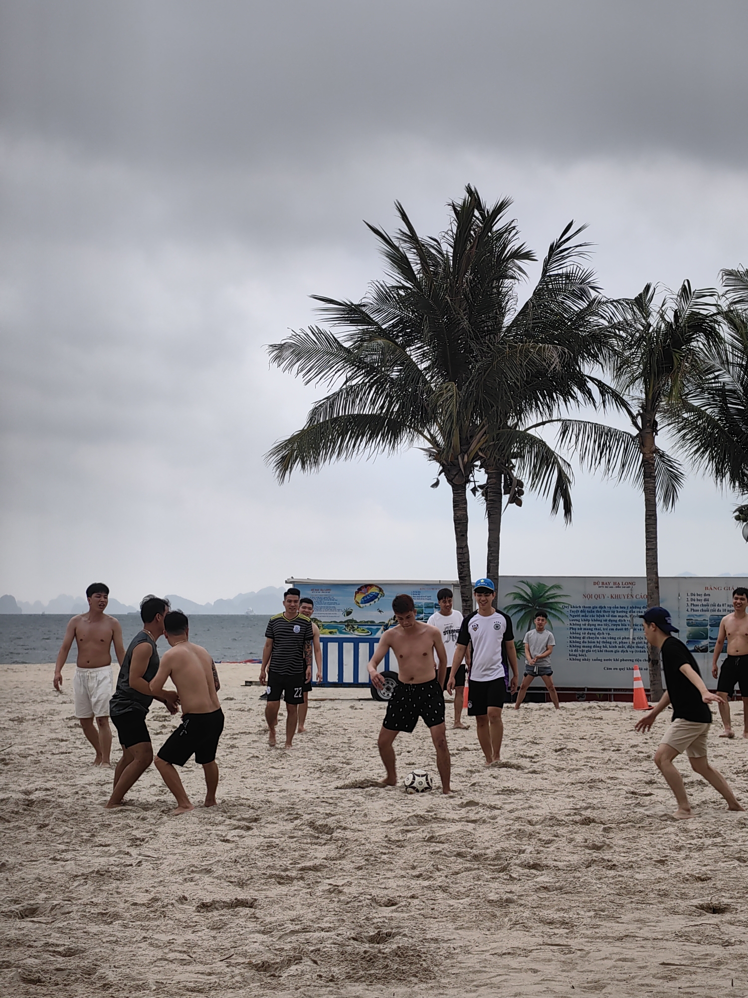 A group of approximately ten men are playing beach soccer on a sandy shore in Bai Chay Ward, Vietnam. Most of the men are barefoot and wearing shorts; some are shirtless, while others wear t-shirts or jerseys. A white and black soccer ball is near the feet of a player in patterned black shorts on the left. The scene is set on a wide, light-brown sandy beach.
In the background, the calm, grey ocean stretches out, meeting a horizon dotted with distant, misty karst islands or mountains, typical of Hạ Long Bay. Several tall palm trees frame the playing area, their fronds reaching towards a uniformly overcast, grey sky, indicating a cloudy daytime.
Behind the players, two large white signs with blue accents are visible. The left sign, titled "DÙ BAY HẠ LONG" (Hạ Long Parasailing), displays "NỘI QUY - KHUYẾN CÁO" (Regulations - Recommendations/Warnings) in Vietnamese. It lists rules for participants, including restrictions for those with certain health conditions, pregnant women, young children, and warnings against drug/alcohol use, entering restricted areas, or bringing valuables. The right sign, labeled "BẢNG GIÁ" (Price List), outlines services such as single and double parasailing, and banana boat rides for groups of 7 or 10 people. The number "22" is visible on the back of one player's jersey, and the word "STRONG" is seen on another's t-shirt. An orange cone acts as a marker on the sand to the right. The overall atmosphere is one of casual recreation on a temperate, possibly humid, cloudy day at the beach.