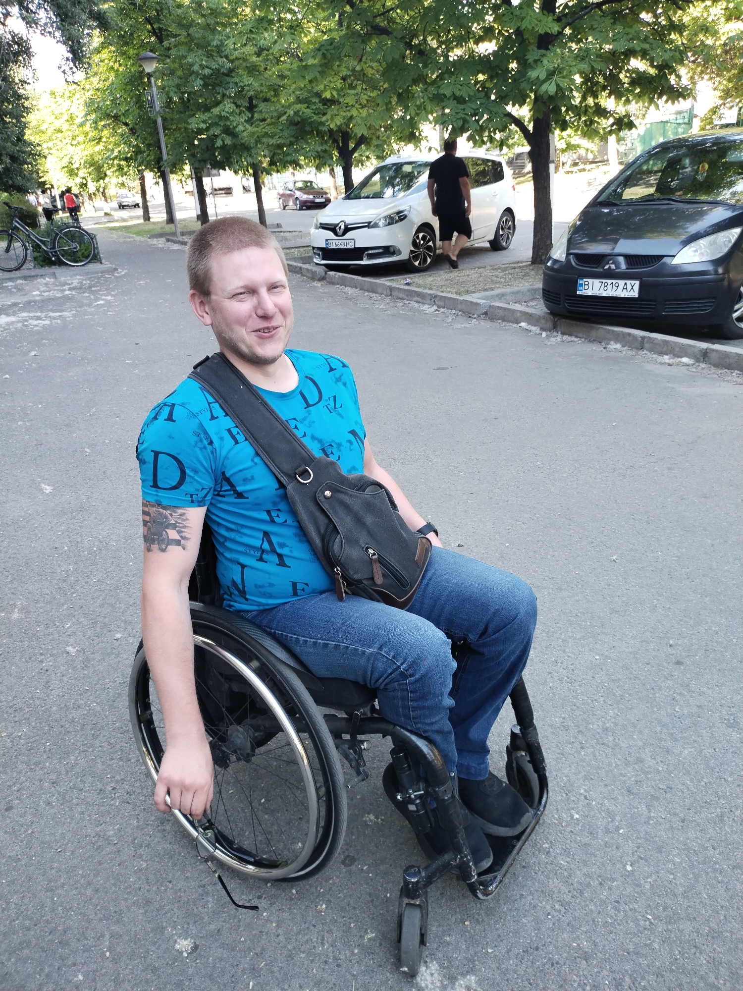 A Caucasian man with short blonde hair is sitting in a wheelchair on an asphalt road in the city of Horishni Plavni, Ukraine. He is wearing a bright blue t-shirt with a repeating pattern of black letters and blue jeans. A black messenger bag with leather accents is slung across his chest. He has a tattoo on his left arm. The man is looking to his left with a smile on his face. The wheelchair has large rear wheels with metal spokes and smaller front wheels. In the background, there are trees lining the street, parked cars, and a lamppost. One car in the background is white with a visible license plate. Another dark gray car is parked on the right side of the image. The scene appears to be a public street or park area.