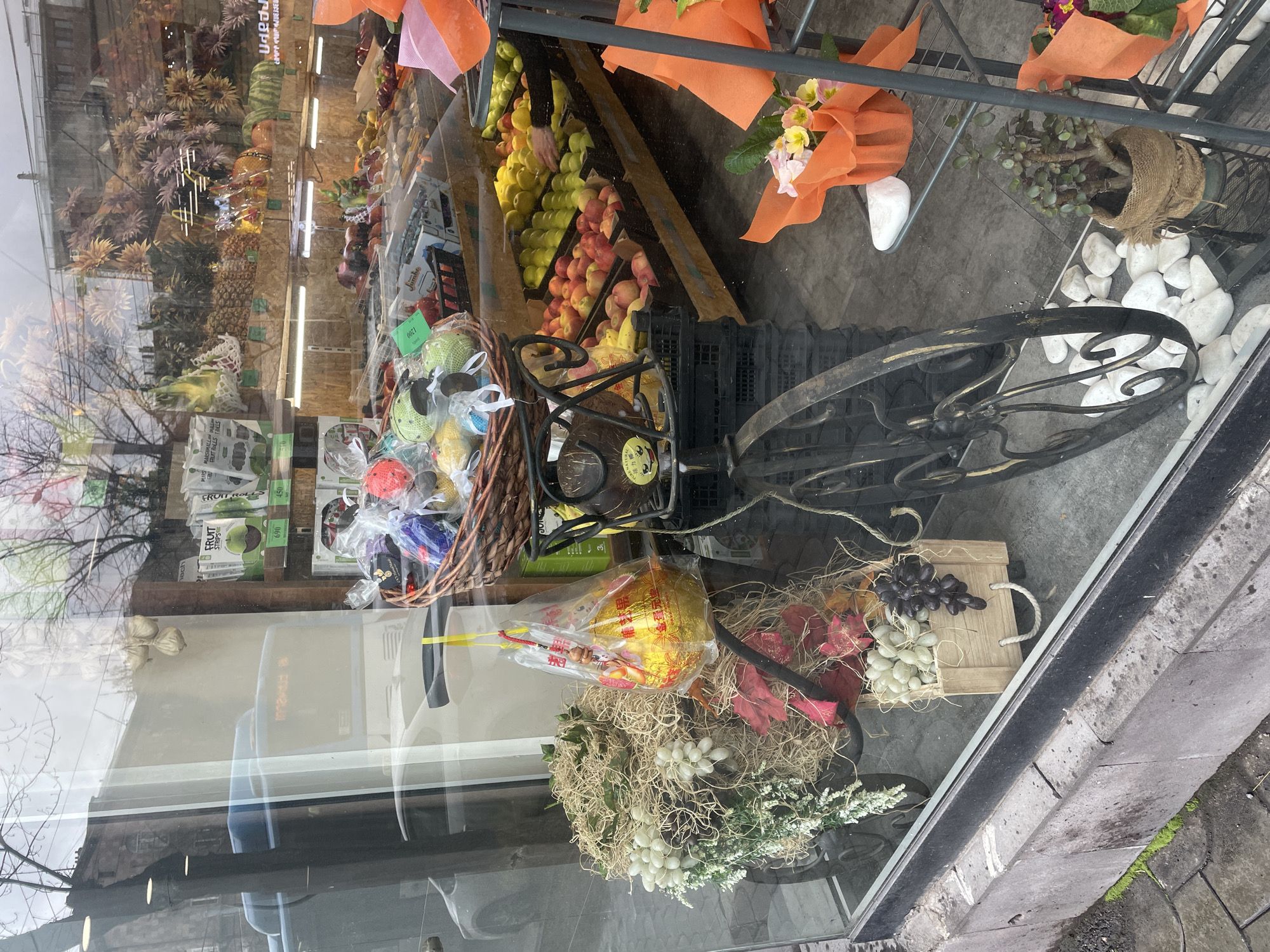 The image presents a view through a large glass display window into the interior of a fruit and grocery store located in Yerevan, Armenia.
Centered within the display is a decorative, stationary black metal bicycle-like structure. The front of this structure is heavily adorned with natural-toned straw, artificial white grapes, and green foliage. A clear plastic bag containing a spherical, gold-wrapped object with red accents is suspended from the handlebar. The rear portion of the bicycle features a basket filled with multiple small, colorful, individually wrapped items, possibly decorative soaps or bath bombs. Below the basket, a small wooden crate contains artificial white and dark grapes. White decorative river rocks are scattered on the floor beneath the bicycle's front wheel.
Inside the store, behind the display, are extensive arrays of fresh fruits. Various types of apples (red, green, yellow) are systematically arranged in black crates on tiered wooden stands, alongside bananas, pineapples, and watermelons. A human hand is visible reaching into a crate of yellow apples. To the left, shelves are stocked with packaged "Fruit Strips," clearly labeled with Armenian text and prices (e.g., 690, 450 Armenian Dram). Dried garlic bulbs are suspended from a wooden beam near these shelves. To the right, metal shelving holds multiple bouquets of cut flowers wrapped in orange paper and a small succulent plant in a burlap-wrapped pot.
Reflections on the glass exterior include an overcast sky, bare tree branches, and the facade of a building. A light-colored vehicle and a blue public bus are also reflected, the bus displaying Armenian text, specifically "ՄԱՐՏԻՆ" (Martiros/Martin), on its destination sign, consistent with routes in Yerevan. The exterior foreground shows a concrete sidewalk composed of grey pavers, with visible patches of moss growth.