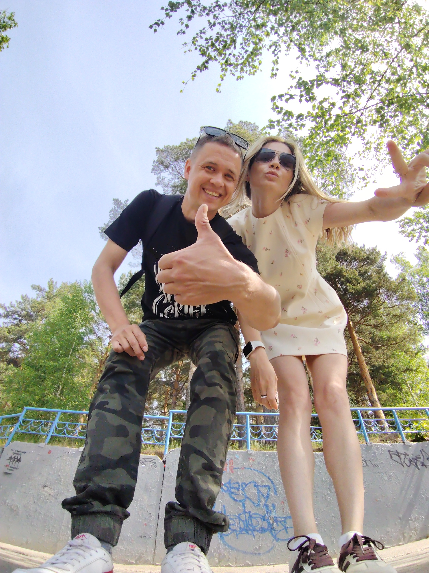 A low-angle shot depicts a man and a woman posing for a photograph outside in Chaikovsky, Russia. The man, dressed in a black t-shirt, camouflage pants, and white sneakers, stands on the left and gives a thumbs-up. Sunglasses rest on his head. The woman, on the right, wears a light-colored dress, sunglasses, and two-tone sneakers; she holds up a peace sign. A blue railing and a concrete wall with graffiti are visible behind them. Trees and foliage fill the background under a sky with scattered branches overhead.