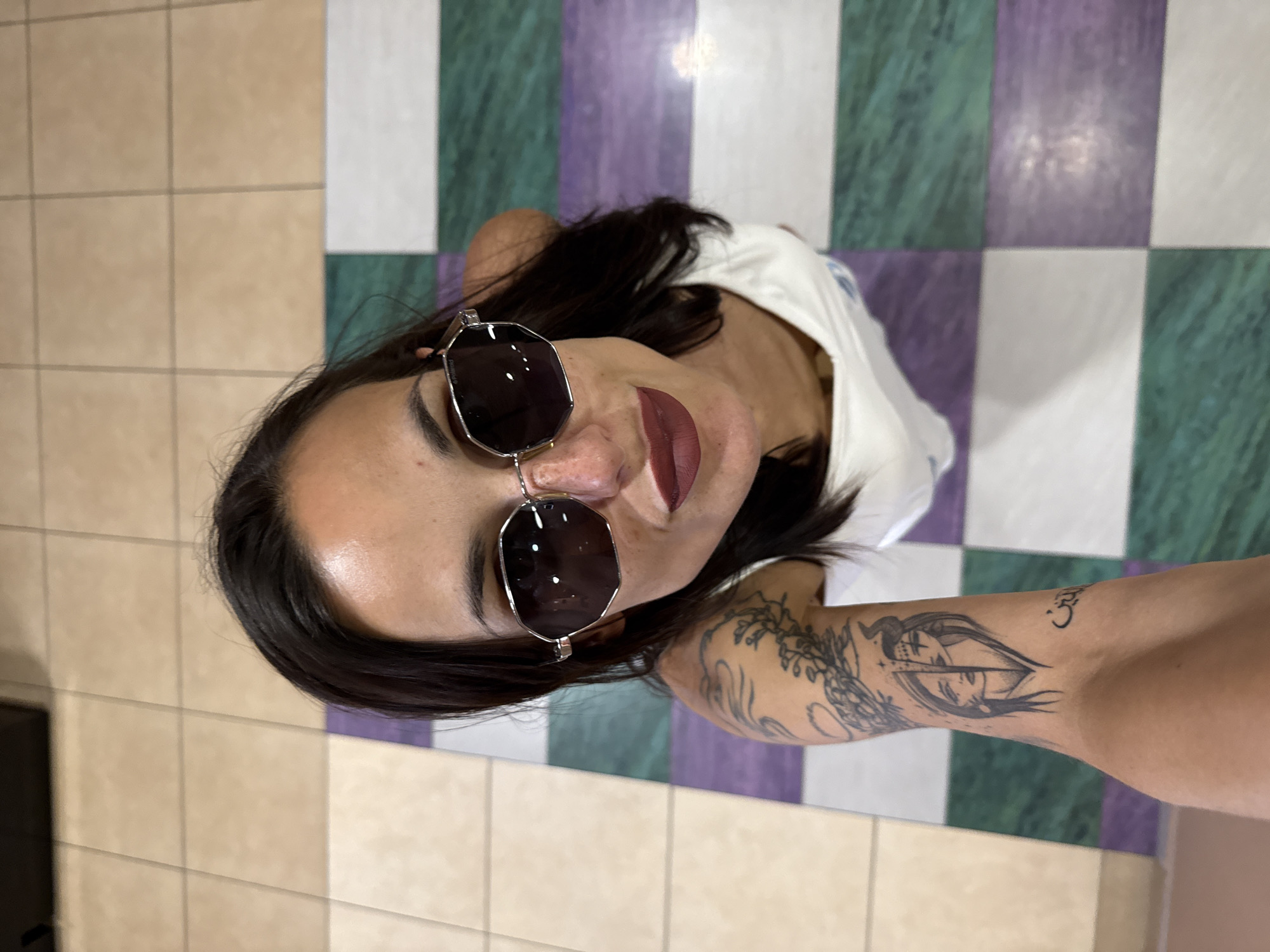 A high-angle, medium shot shows a woman with dark hair and sunglasses looking up at the camera, with a tattoo on her left arm. She is wearing a white top. The background consists of a tiled floor with alternating beige, purple, and green squares. The image appears to have been taken indoors, possibly in the city of Pleven, Bulgaria, given the context of previous interactions.