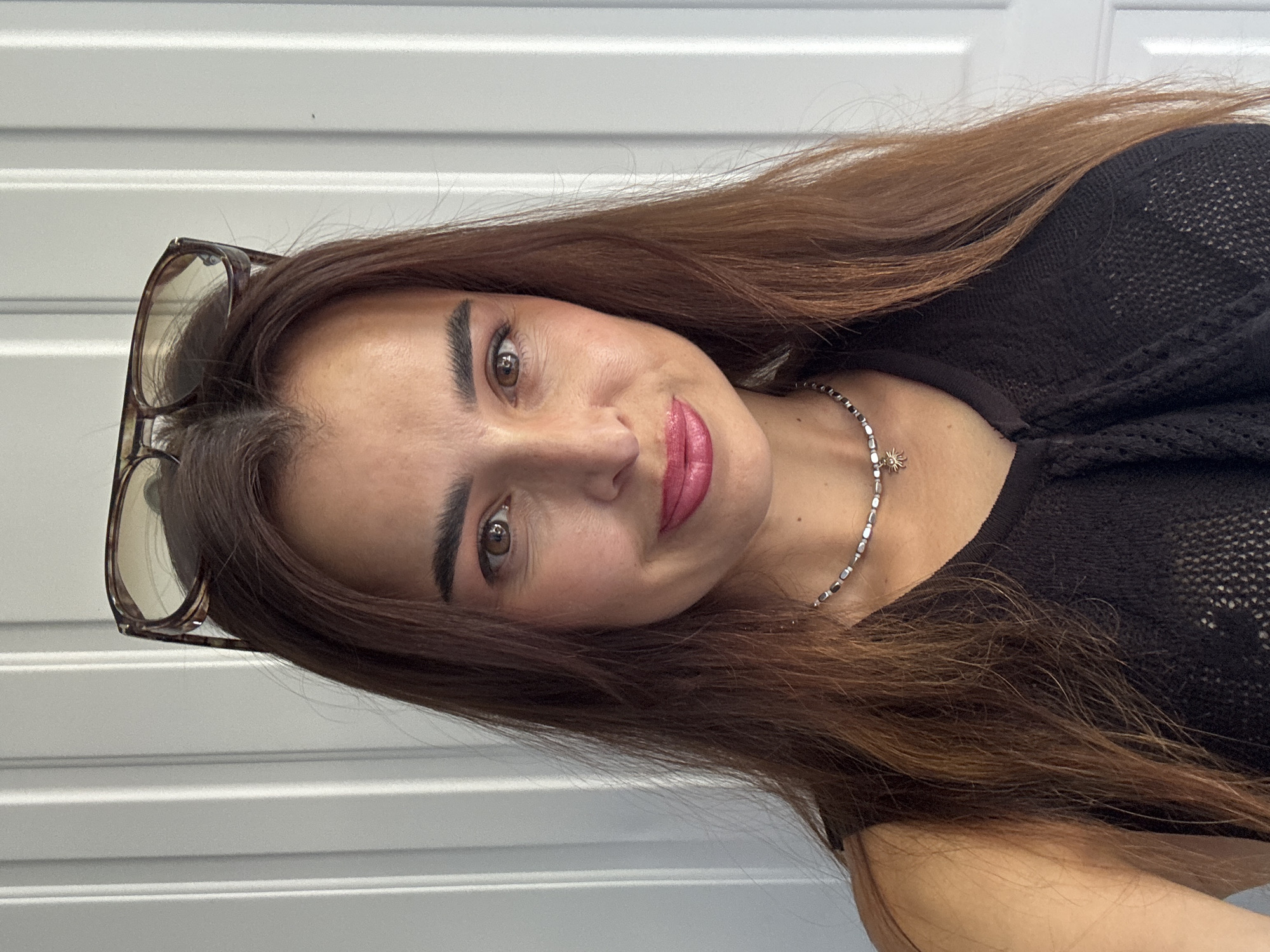 A medium close-up selfie features a woman with long, reddish-brown hair, wearing a black, open-knit sleeveless top and a silver beaded necklace with a small sun charm. Sunglasses are perched on top of her head. Her eyes are brown, and she is wearing makeup, including pink lipstick. The background is a vertical ribbed white surface, possibly a door or wall. The lighting appears to be natural, casting subtle shadows. The image is sharp and focused on the woman's face and upper body.