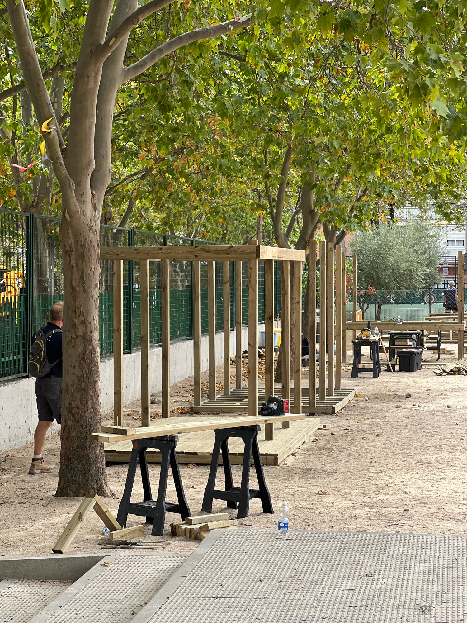 The image depicts an outdoor construction site. In the foreground, there are two black sawhorses supporting wooden planks. Scattered on the sandy ground are pieces of lumber and construction debris. In the midground, a partially constructed wooden structure is visible, with vertical posts and horizontal beams. A person wearing a backpack and shorts stands near a large tree, facing away from the camera. Behind the construction, a green metal fence runs along the perimeter, with a concrete wall beneath it. A yellow piece of machinery is partially visible beyond the fence. Further in the background, trees with green foliage are present, along with a glimpse of buildings and a tennis court. The scene appears to be an open-air area, possibly a park or recreational space undergoing renovation or construction. The lighting suggests it is daytime, with the sun casting shadows. This image was taken in Getafe, Spain.