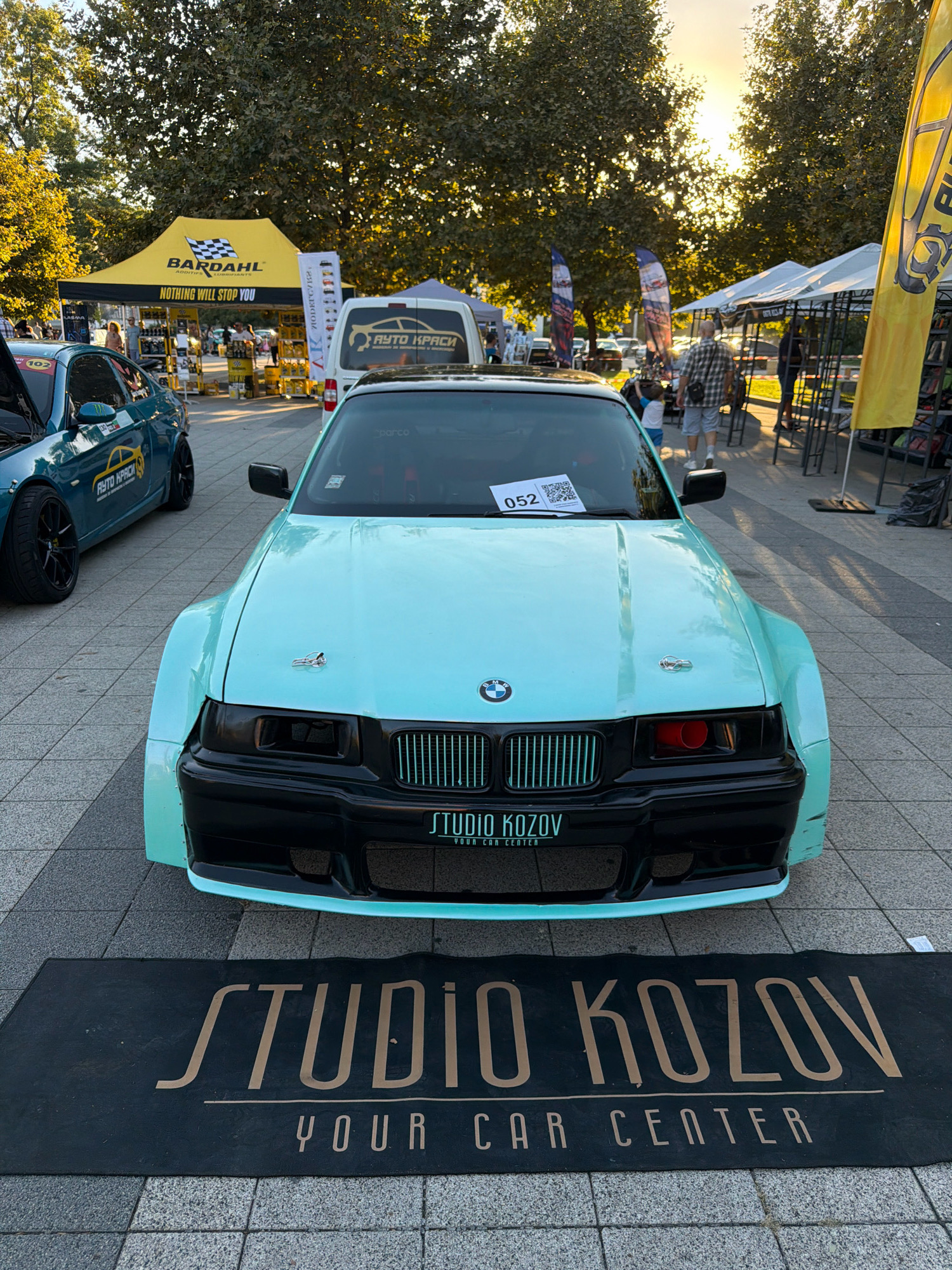 The image depicts an outdoor event, possibly a car show, in Pleven, Bulgaria. In the foreground, a light blue BMW E36 with a widebody kit and black accents is parked on a paved area. The car has a visible license plate or sticker with "052" on the windshield. A black mat with the text "STUDIO KOZOV YOUR CAR CENTER" is placed in front of the car.
Behind the BMW, a teal-colored BMW E92 is visible with its hood open, also displaying "AUTO HRACİ" branding. In the background, there are several tents, including a yellow "BARDAHL" tent with "NOTHING WILL STOP YOU" branding. Various promotional banners and displays are set up. People are seen walking around the event, including a man in a plaid shirt and a child in a blue shirt walking away from the camera. The sun is setting, casting a warm glow and creating lens flare through the trees. The overall atmosphere suggests a community gathering focused on automotive culture.