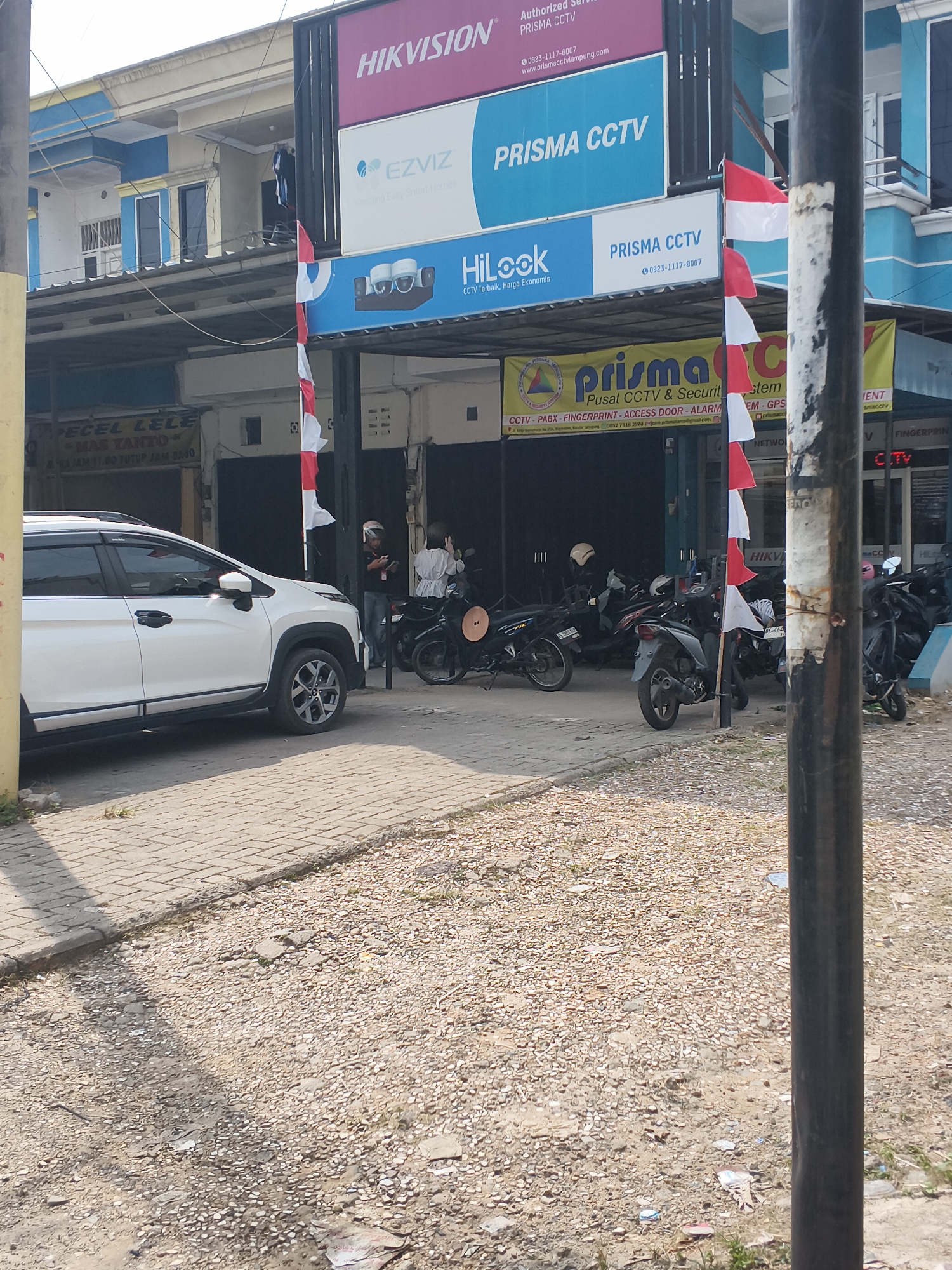 The image captures a street scene in Bandar Lampung, Indonesia, dominated by a storefront and its signage. The storefront appears to be a business specializing in CCTV and security systems, with prominent banners displaying brands like HIKVISION, EZVIZ, and HiLook. A company named "Prisma CCTV" is heavily advertised, with their contact number (0823-1111-8007) visible on multiple signs. The business also seems to offer services related to PABX, fingerprint access, and alarm systems, as indicated by a banner stating "Pusat CCTV & Security Sistem".
In front of the store, a white SUV is parked on a paved sidewalk, partially obscuring the entrance. Several motorcycles are also parked in front of the store, suggesting it is a busy commercial area. Two individuals, possibly employees or customers, are seen near the motorcycles, one of whom is looking at a mobile phone. The overall environment suggests a typical urban commercial strip with a focus on security technology retail. Indonesian flags, red and white, are displayed on poles near the entrance, indicating a celebration or national observance. The ground in front of the paved area is covered with gravel and debris.