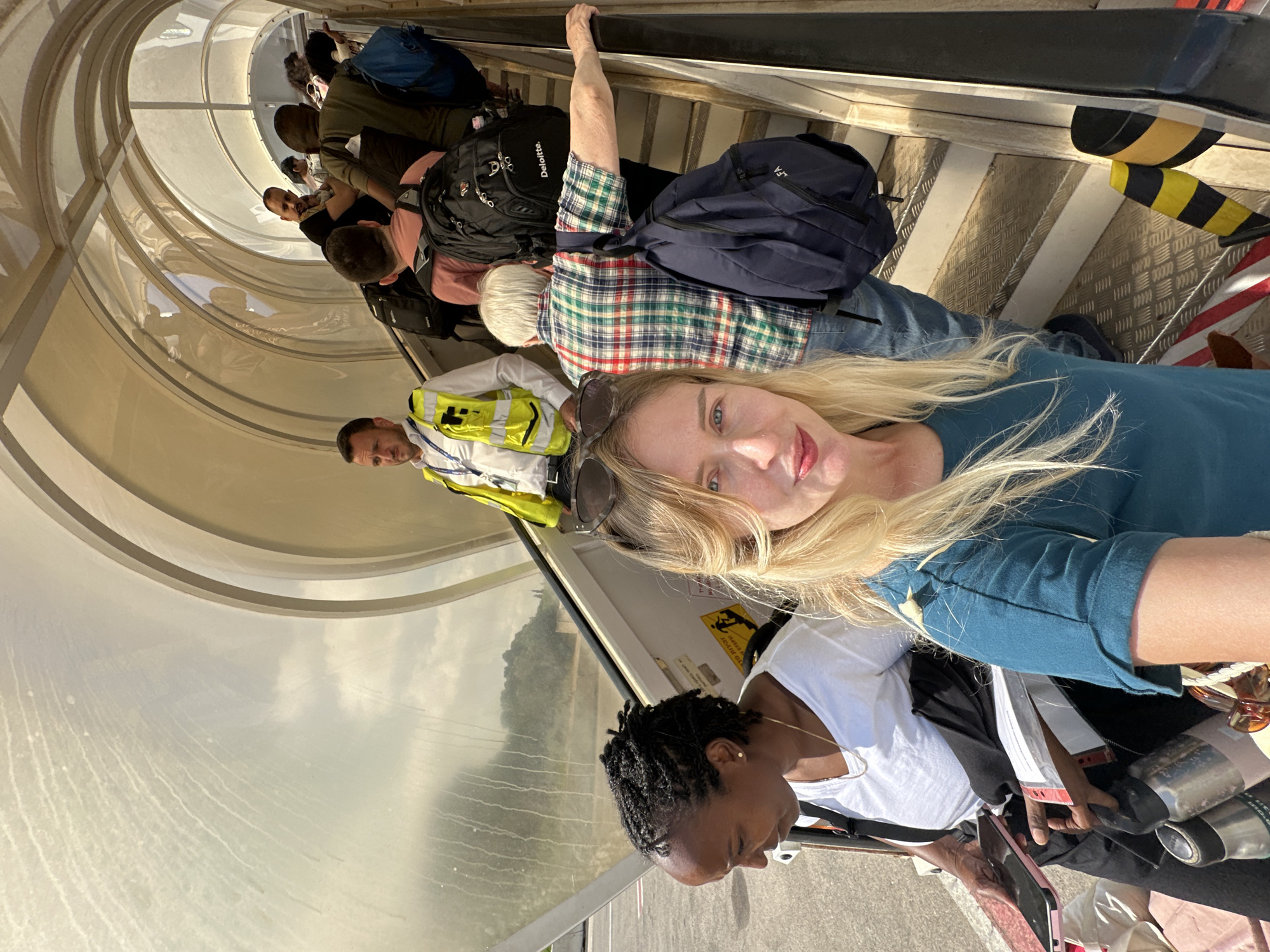 In Yehud-Monosson, Israel, people are boarding a plane via an outdoor staircase. The image features a woman in the foreground taking a selfie, with another woman walking up the stairs to her left. A man in a yellow vest is visible further up the stairs, seemingly directing passengers. The environment appears to be an airport tarmac under a cloudy sky, with a covered walkway leading up to the aircraft. Passengers are carrying luggage, including backpacks.