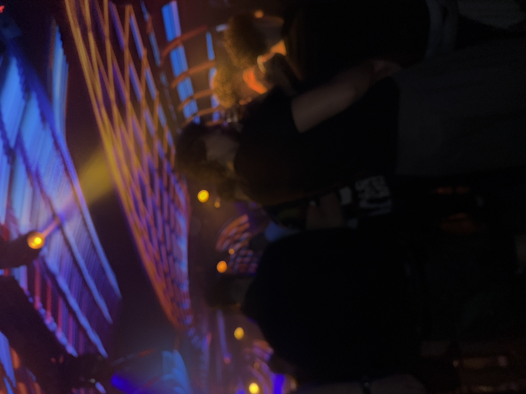 The image shows a blurry scene with illuminated structures or decorations. The dominant colors are blue, orange, and yellow, suggesting the presence of lights. There are individuals in the scene, their details obscured by blur and darkness. The environment appears to be outdoors, potentially at night, in the city of Unknown, Unknown, due to the blurry lights and figures. The lack of clarity makes a more precise assessment impossible.