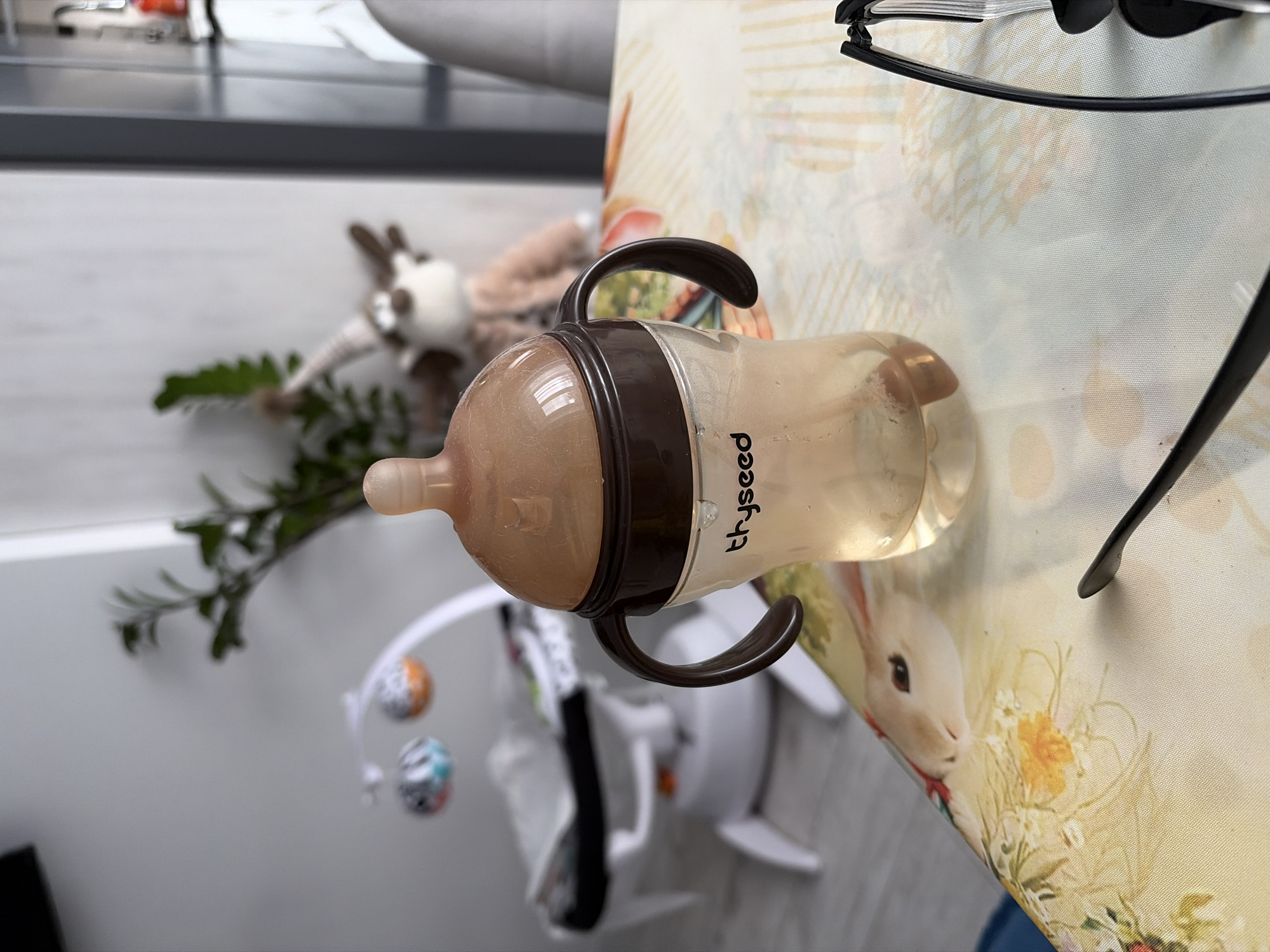 The image shows a baby bottle with brown handles and a nipple, lying on a table with a patterned tablecloth featuring images of rabbits and flowers. The bottle contains a light-colored liquid and has the word "tryseed" printed on its side. In the background, a plush animal, possibly a deer or moose, and a baby mobile with colorful balls are visible. A pair of glasses is also present on the table next to the bottle. The location is not discernible from the given image.