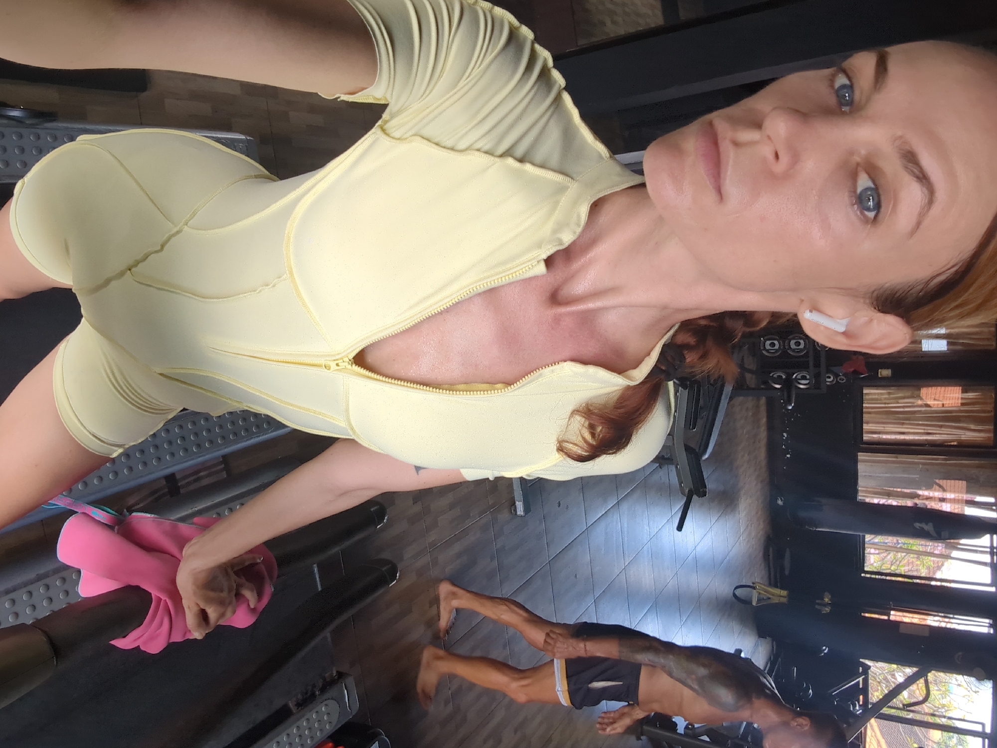 The image shows a woman in a yellow bodysuit at a gym, perspiring, with an earbud in her right ear. She holds a pink towel near a treadmill. In the background, another person is exercising. The location is a gym in Mengwi, Indonesia.