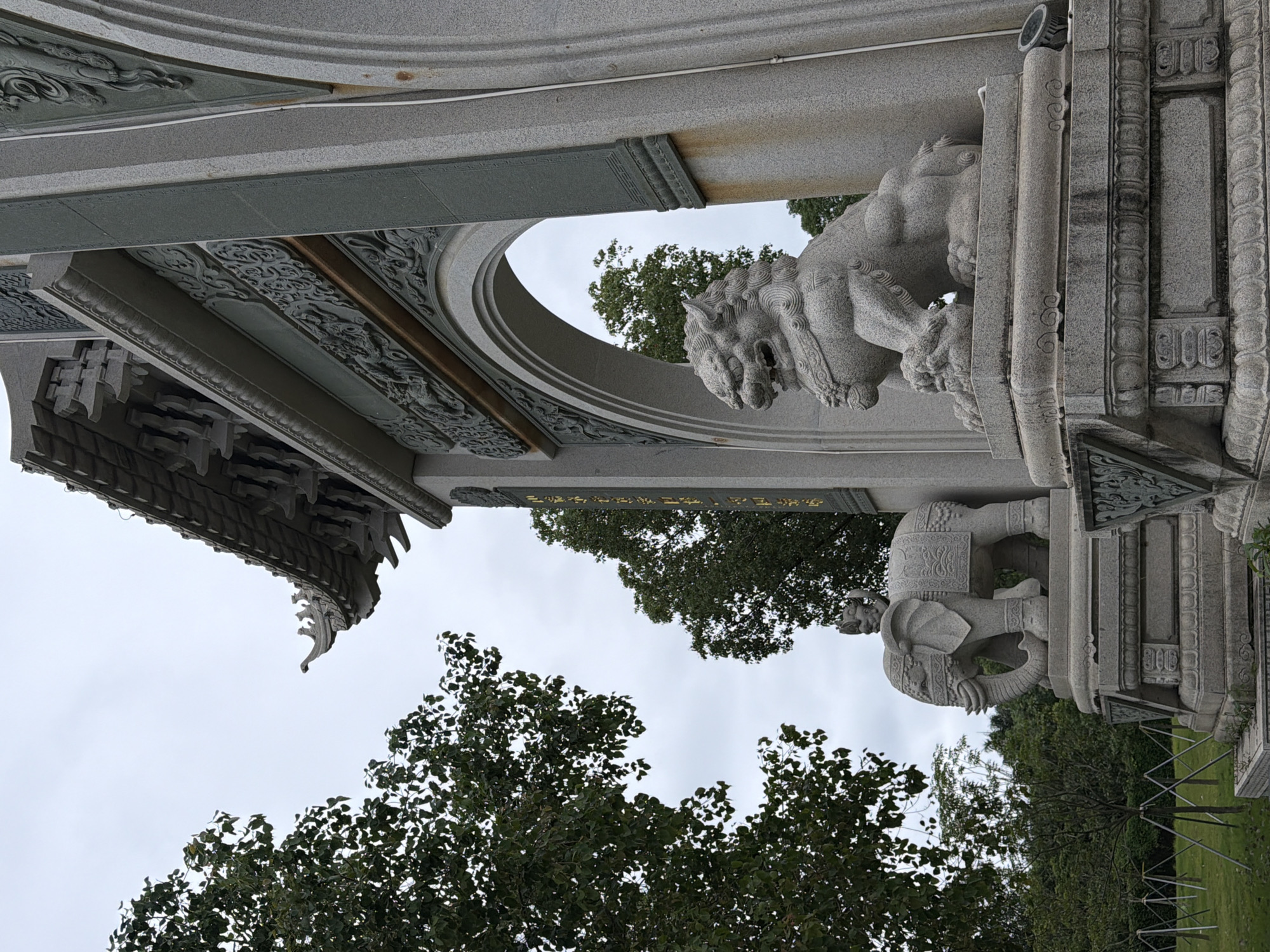 The image depicts a stone guardian lion statue in front of an ornate gateway, likely in Longgang District, China. The lion is positioned on a stone pedestal, its body in a seated posture with its mouth open and mane flowing. To the left of the gateway, a stone elephant statue with a rider is visible, also on a pedestal. The gateway itself is constructed of grey stone, featuring intricate carvings along its top and sides. The roof of the gateway has a distinctive tiered design with decorative elements. Green trees with dense foliage surround the area, and the sky above is overcast. The overall scene suggests a cultural or religious site, possibly a temple or a memorial.