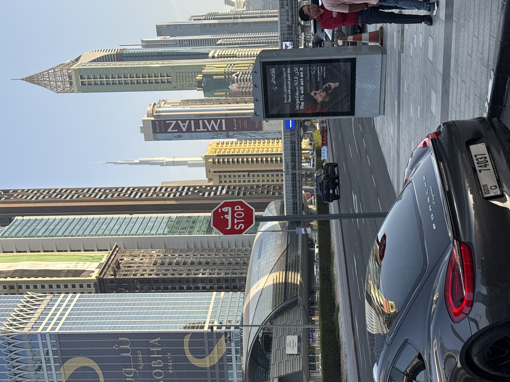 The image captures a street scene in Dubai, United Arab Emirates, likely in the Downtown Dubai or Business Bay area given the prominent skyscrapers. In the foreground, the rear of a dark grey Porsche Cayman is visible, with its license plate reading "74037 Q". To the right of the car, two men are standing on a sidewalk. One man is wearing a red shirt and black pants, and the other is wearing a light pink shirt and blue jeans. Beside them is a digital billboard displaying an advertisement for "Shuraa", with text in Arabic and English stating "Most will scroll past this but the 1% will act on it. Your business in Dubai starts here." In the background, a modern metro station with a curved, metallic roof is visible, along with numerous tall, modern buildings characteristic of Dubai's skyline, including a glimpse of the Burj Khalifa in the distance. A red octagonal stop sign with Arabic text is also present. The scene appears to be illuminated by natural daylight, suggesting it was taken during the daytime.