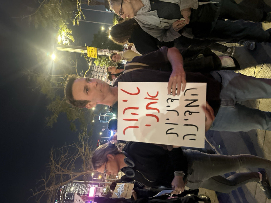 The image captures a nighttime scene of a public gathering or protest, oriented vertically with people appearing to stand sideways in the frame. In the immediate foreground, a man with short, dark hair and wearing a dark top holds up a white protest sign. He looks directly at the camera with a neutral to slightly serious expression.
The sign he holds displays text in Hebrew, written in two columns. The right column, in black text, reads "המדיניות" (Ha-mediniyut), which translates to "The policy." The left column, in red text, reads "הורגת אותנו" (Horeget otanu), which translates to "is killing us" or "kills us." Together, the sign reads: "המדיניות הורגת אותנו" ("The policy is killing us").
Behind the man, other individuals are visible, suggesting a crowd. To the man's left (viewer's right, if the image were rotated correctly), a woman with short, light-colored hair and glasses is looking downwards. She has a pair of headphones resting around her neck. Further back, another woman with darker, curly hair is partially visible. The faces of other people in the background are mostly obscured or blurred.
The setting is outdoors, likely a city street or plaza, illuminated by bright artificial streetlights that create a strong glow and some lens flare in the upper left of the frame. The sky is dark, indicating it is night. In the deeper background, there are outlines of urban buildings and trees. A yellow street sign with some indistinct Hebrew text is visible in the distance, illuminated by the streetlights. The overall atmosphere suggests a demonstration or rally taking place after dark.