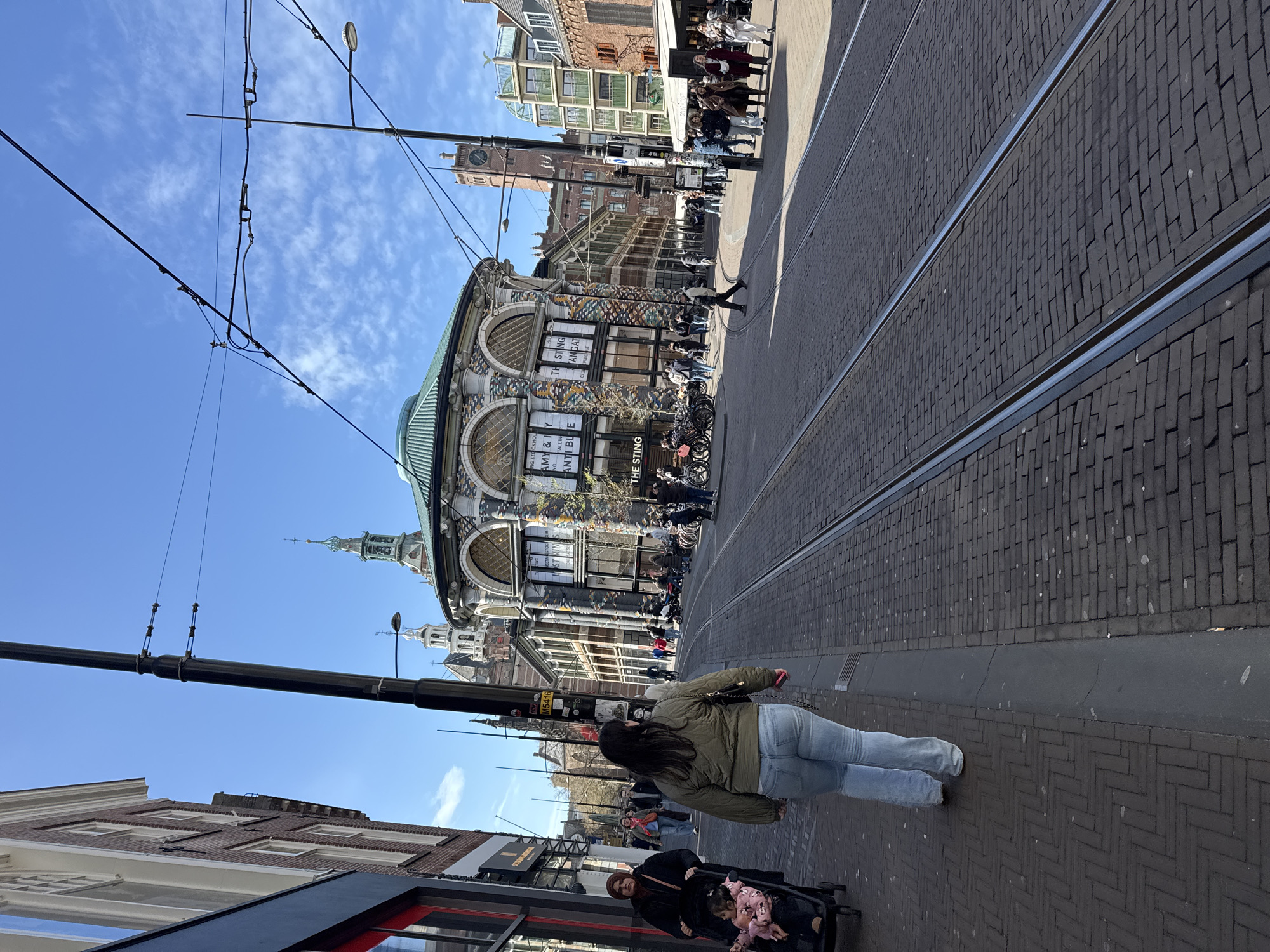 The image captures a street scene in The Hague, Netherlands, identifiable by the presence of tram tracks embedded in the brick-paved road. A woman in a green jacket and jeans walks along the sidewalk, away from the camera. To the lower right, another woman with a stroller is visible. The architectural style of the buildings suggests a historic European city center. The prominent building in the background features a distinctive dome and decorative facade. Overhead tram lines traverse the scene, and streetlights line the road. Several people are visible in the background, along the storefronts. A sign partially visible indicates a store named "The Sting".