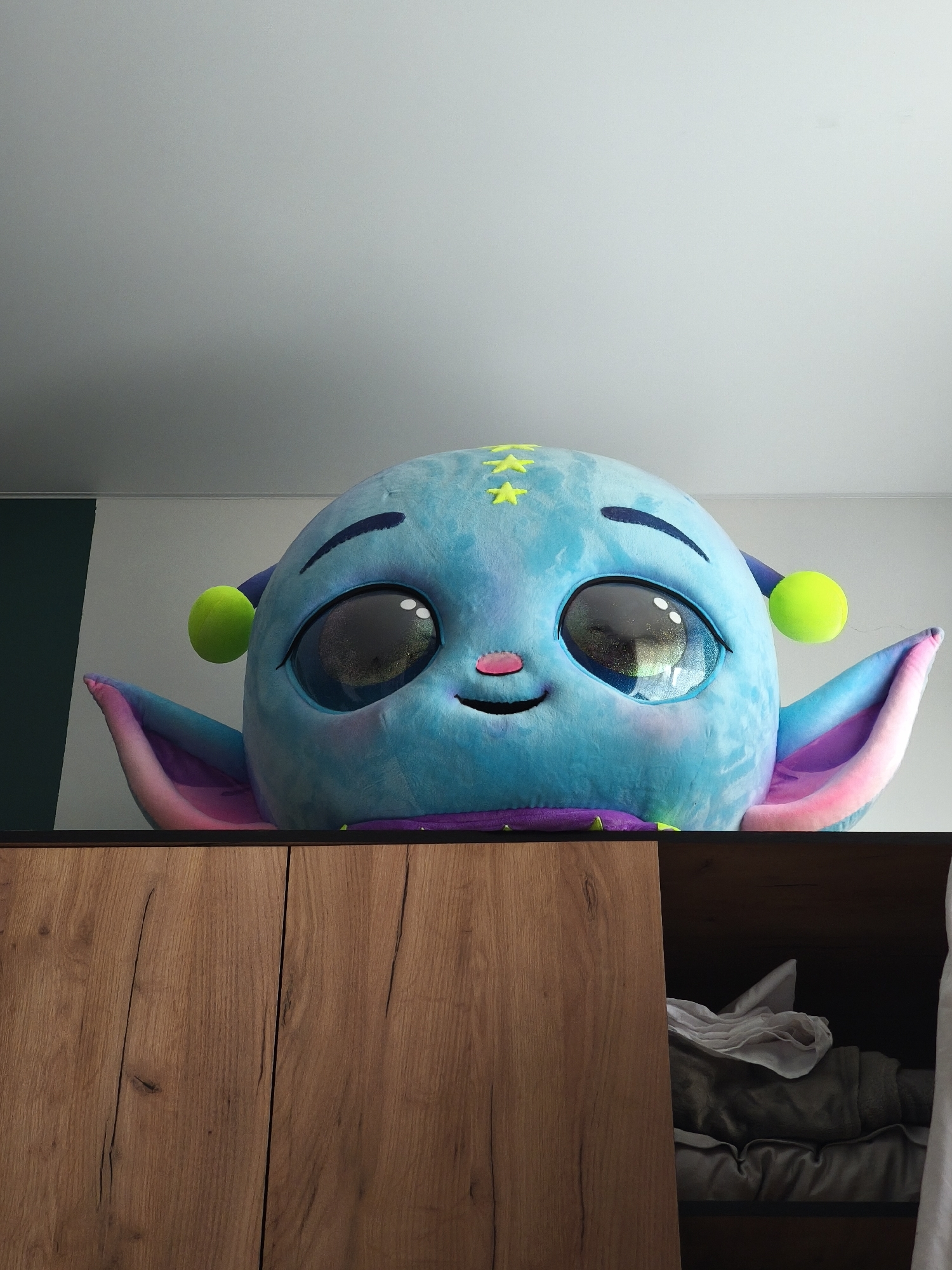A large, blue, plush, alien-like toy with green ear ornaments and star-shaped decorations on its head is peeking over the top of a wooden cabinet. The toy has large, reflective eyes, a pink nose, and a smiling expression. The interior of the partially open cabinet is visible, revealing folded clothing and linens. The background consists of a white wall and a vertical green panel. The location is not specified, and there is no indication that the image was taken in Moscow, Russia.