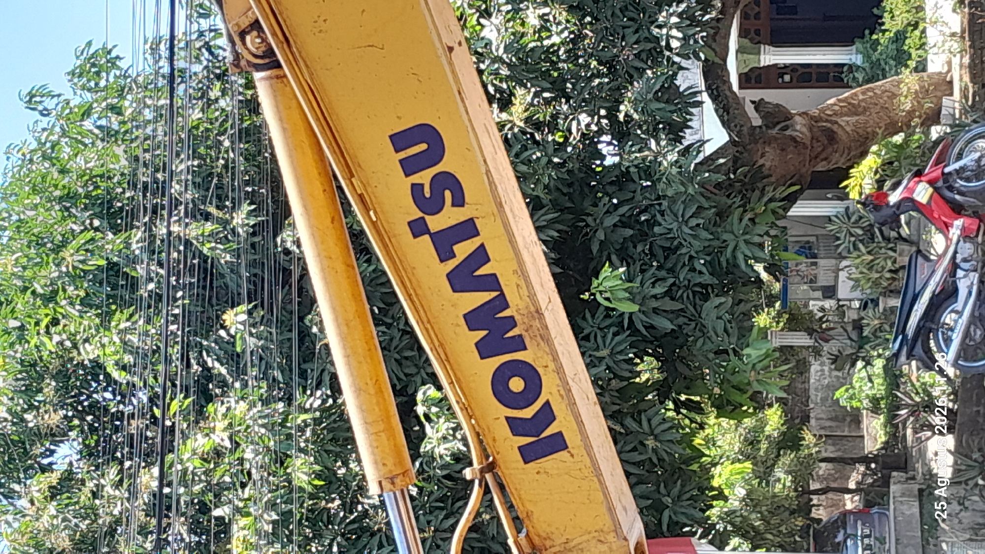 The image shows a close-up of a yellow construction vehicle arm with the blue text "KOMATSU" visible on it. The arm is angled upwards, with a hydraulic cylinder at the top left. Behind the arm is a dense green tree canopy, with power lines visible above it. The bottom of the image shows more of the tree, as well as part of a house and a red motorcycle. The bottom left corner of the image contains a timestamp: "25 Agustus 2026 11:26". The scene appears to be outdoors, likely in a residential area in Jomblang, Indonesia, given the presence of the house, motorcycle, and lush vegetation common in tropical regions. No people are visible in the frame.
