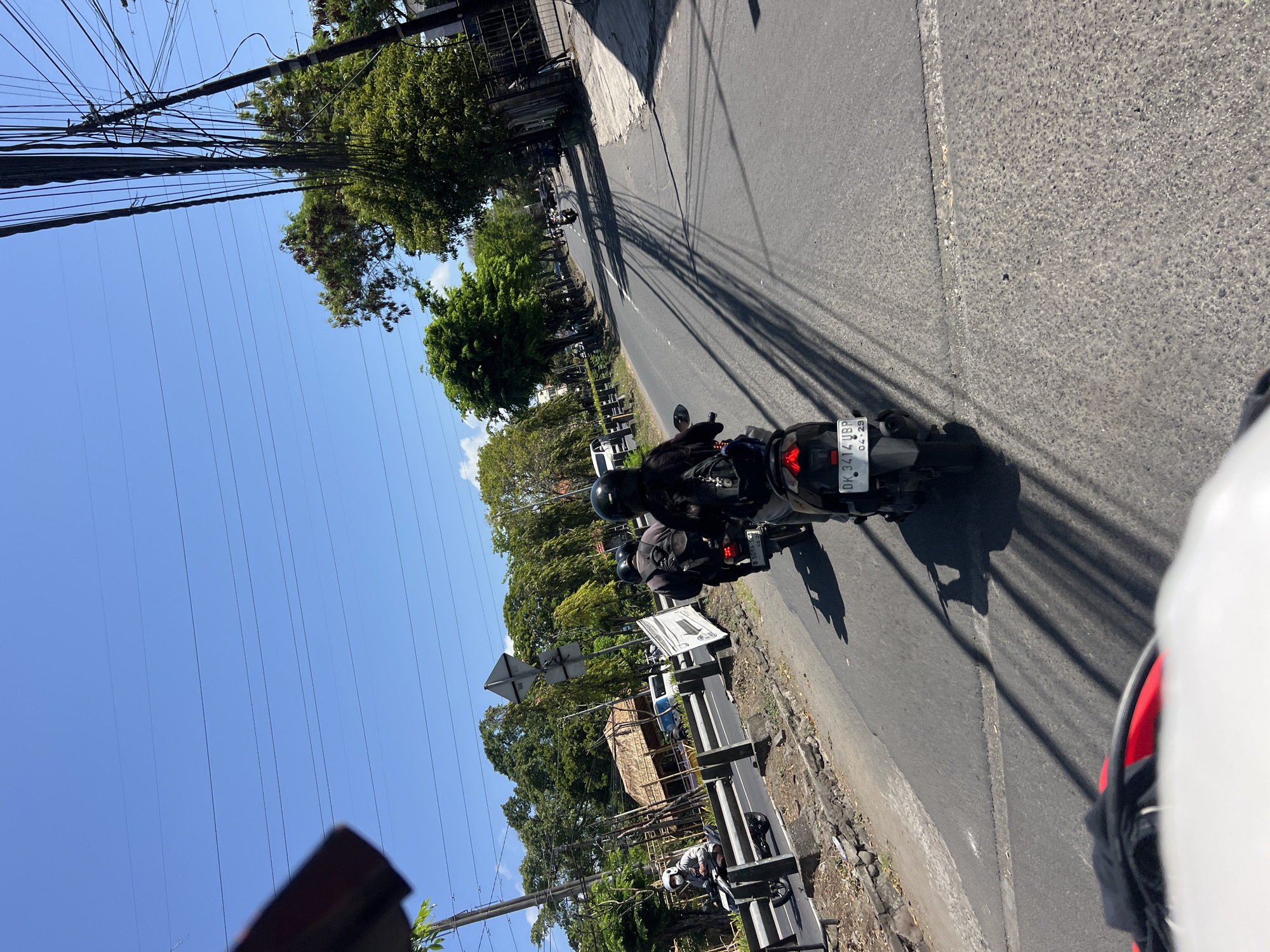 The image depicts a sunny day in Denpasar, Indonesia, with clear blue skies and scattered clouds. The scene is captured from the perspective of someone riding a motorcycle, showing the road ahead and the surrounding environment. Several motorcycles are on the road, with riders wearing helmets. On the left side of the road, there are trees, a traditional thatched-roof building, and a white van. Utility poles with numerous wires are visible on the right side of the road, casting long shadows across the asphalt. The road itself is paved and appears to be a main thoroughfare in the city. The overall impression is one of a typical day in a tropical urban setting.
