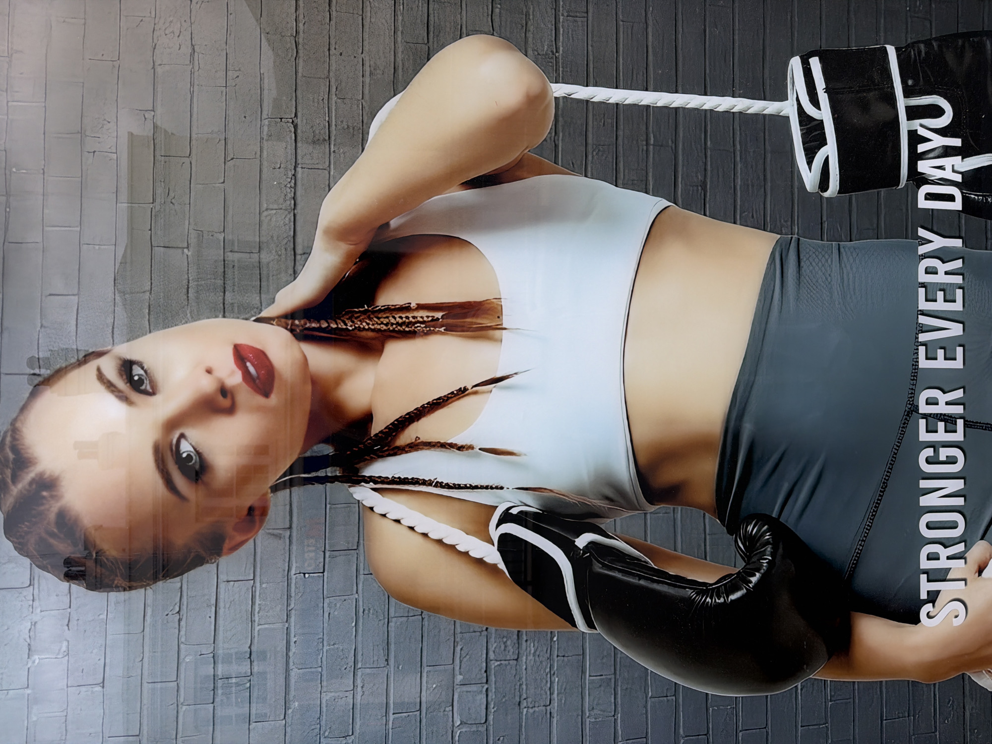 The image shows a poster of a woman in athletic wear and boxing gloves. She is wearing a light blue sports bra and gray leggings, and her hair is styled in braids. She is standing in a boxing ring, with the ropes and corner post visible. The text "STRONGER EVERY DAY" is vertically displayed along the right side of the image. The background is a gray brick wall.
Without additional information, I cannot confirm the location in Epsom and Ewell, United Kingdom.