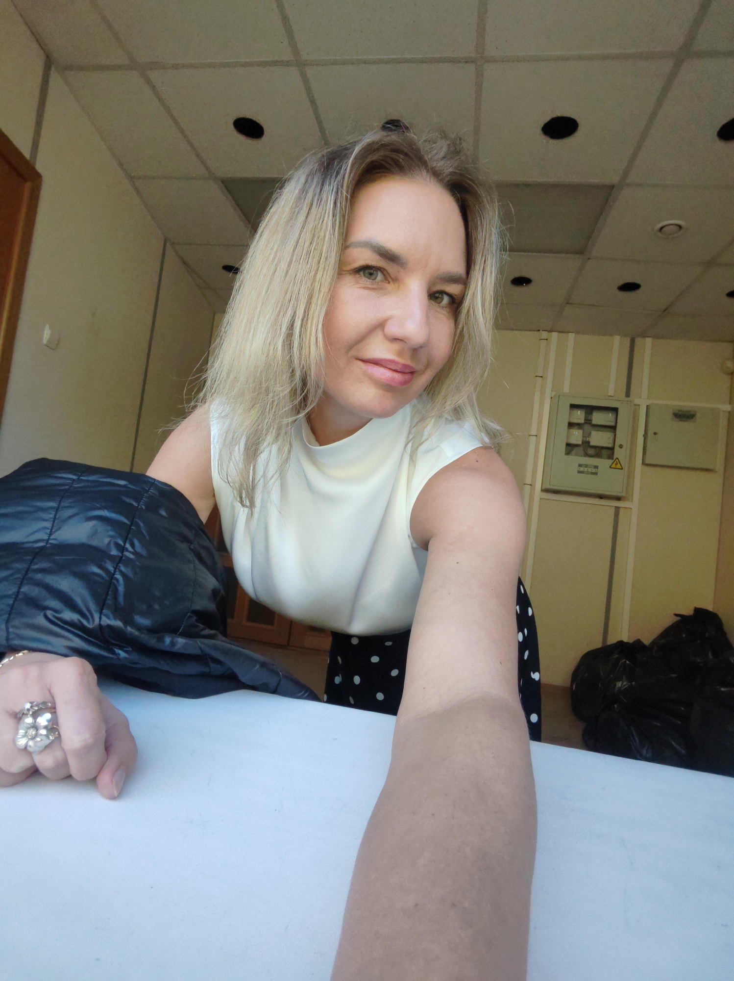 A woman with blonde hair and green eyes takes a selfie indoors. She is wearing a white sleeveless top and a black skirt with white polka dots. She is leaning on a white surface, and a black jacket is draped over her left arm. Her right hand is visible in the foreground, wearing a large ring with a flower design. In the background, there is a suspended ceiling with lights, a wall with electrical panels, and two black garbage bags. The environment appears to be an office or utility space.