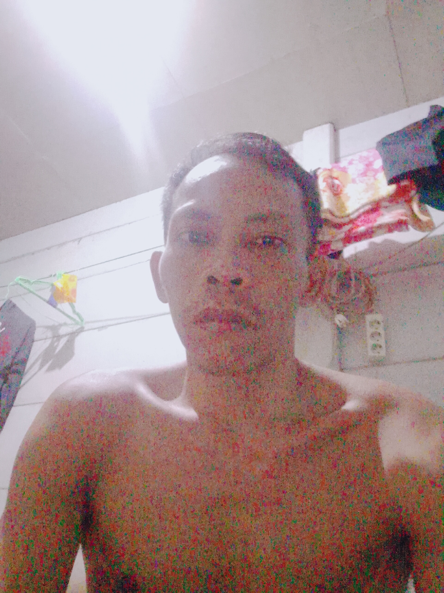 A close-up, slightly high-angle photograph depicts a shirtless man with short, dark hair. The man is facing forward, his head and shoulders filling much of the frame. His skin appears reddish due to the photograph's visual noise.
Behind the man is a room with white, horizontally-paneled walls. On the left, there is a clothes hanger with some clothing items. In the upper right corner, there are folded towels or blankets. An electrical outlet is visible to the right of the man's head.
The lighting in the room is bright, casting a glare from above, and appears to originate from an interior light source. The photograph's resolution is low, with considerable digital noise. The location is Bukit Ketok, Indonesia, which is relevant only as it is given context.