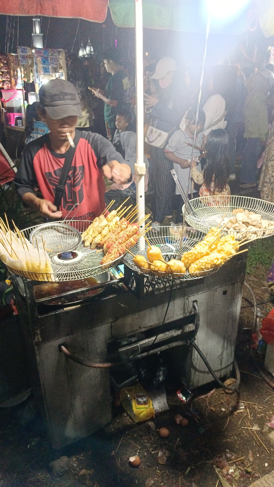 In Tajurhalang, Indonesia, a street food vendor is preparing food at a night market. The vendor, wearing a black cap and a red and black t-shirt with a Transformer logo, is seen holding a cigarette and attending to a food cart. The cart features a metal grate filled with various fried skewers, including some coated in batter and others that appear to be sausages. Another grate holds what seem to be fried meatballs or tofu. The cooking apparatus itself is a metal box with a visible exhaust pipe. The surrounding environment is dimly lit, typical of a night market, with a crowd of people visible in the background, suggesting active commerce. There are also some discarded eggshells and debris on the ground near the cart, indicating an outdoor, possibly informal, setting.