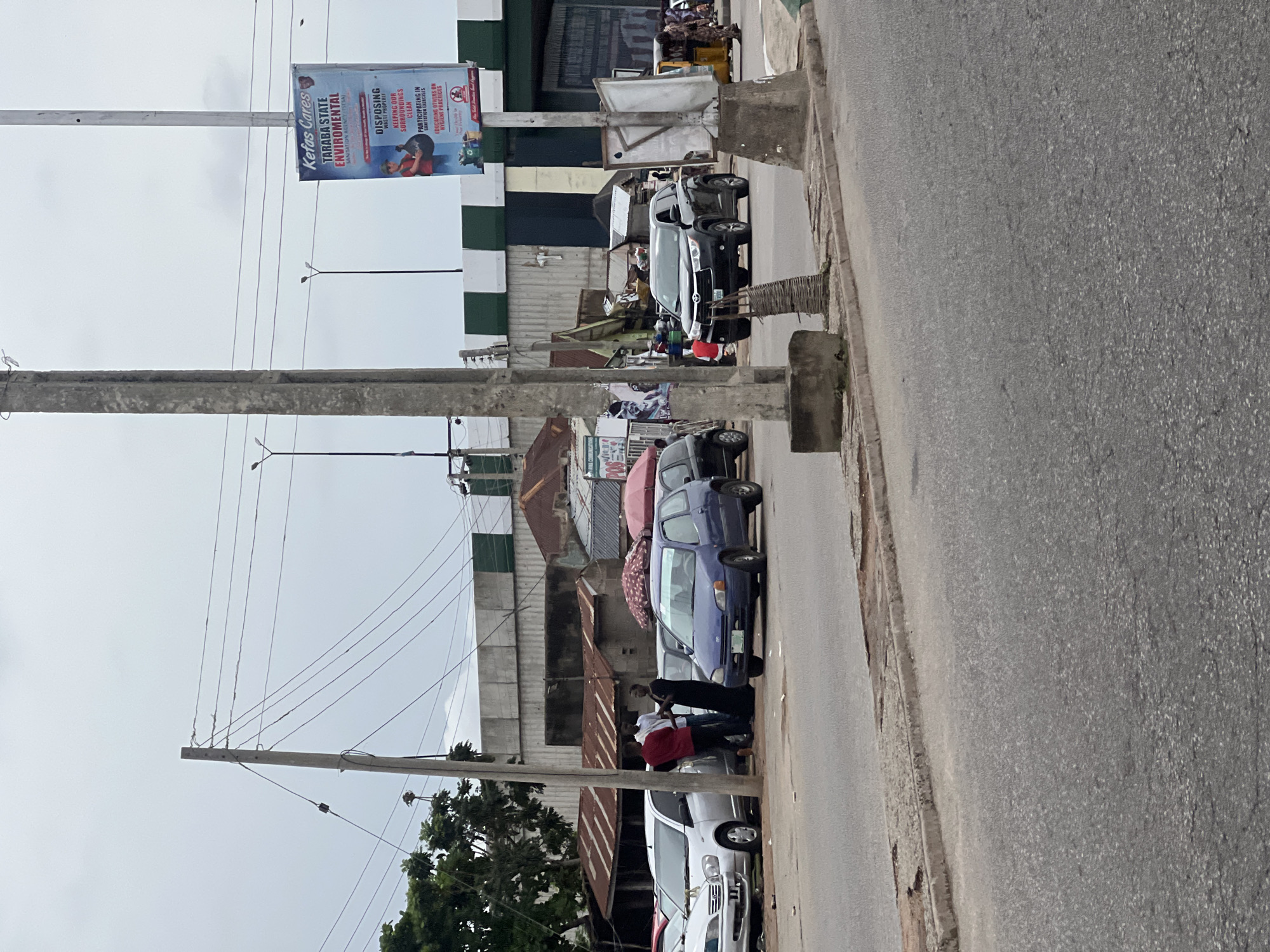 The image depicts a street scene in Jalingo, Nigeria, under an overcast sky. In the foreground, a paved road transitions to a curb and sidewalk. Several vehicles are present: a silver sedan is parked on the left, a blue hatchback is in the center, and a black SUV is on the right, in motion. A yellow auto-rickshaw is visible further down the road on the right.
A group of people are gathered near the silver sedan, some engaged in conversation. Various structures, including corrugated metal-roofed buildings and a large billboard, line the street. The billboard, located on the right side of the image, displays text in English and an image related to environmental sanitation, featuring the logo "Kefas Cares Taraba State Environmental." Electrical poles and wires crisscross the upper portion of the scene. Trees are visible on the far left. The overall impression is of a bustling urban environment with everyday activities taking place.