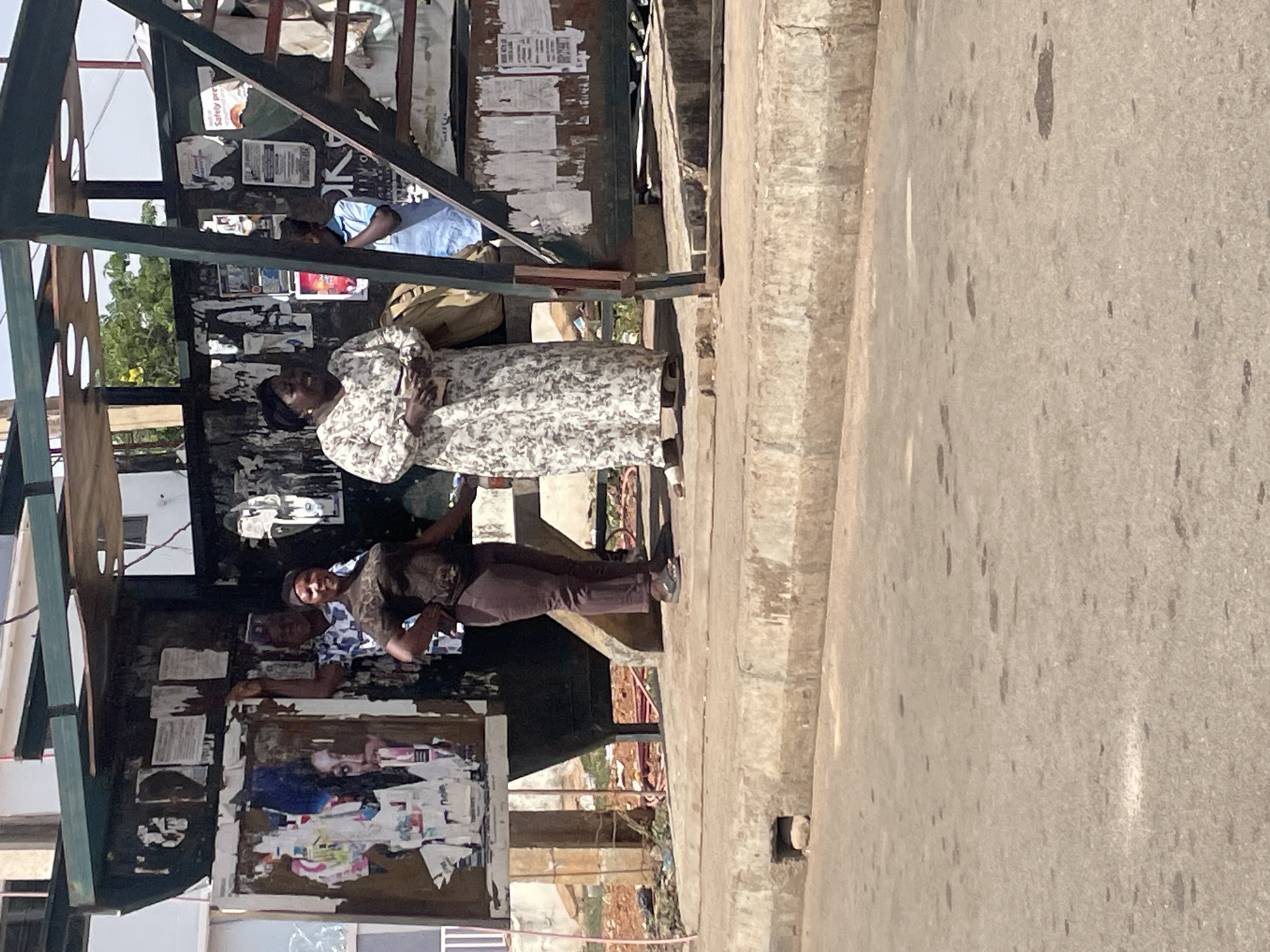 The image shows a street scene, likely in the city of Jos, Nigeria, based on visual cues. A woman in a white and floral dress is standing under a metal structure, possibly a bus stop or market stall, with posters and notices attached to its frame. Behind her, two men are seated within the structure. The man closer to the camera is wearing a printed shirt and brown pants. To the right of the structure, a concrete sidewalk and road surface are visible.