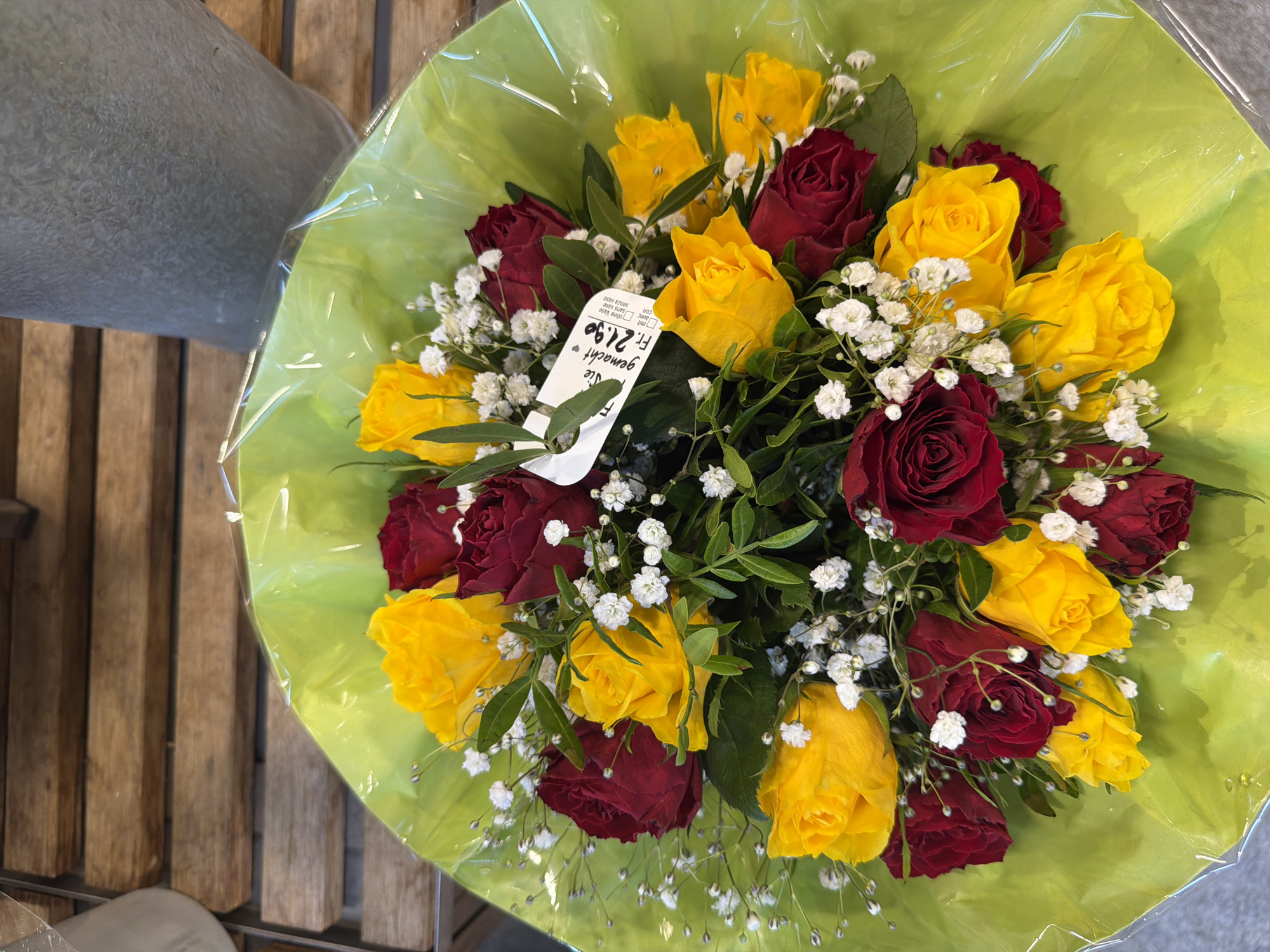 A bouquet of red and yellow roses is presented in clear plastic wrapping. The bouquet is filled with small white flowers and green leaves. A white tag is attached to the bouquet with the text "Fr. 21.90" printed on it. The bouquet is placed on a wooden surface.