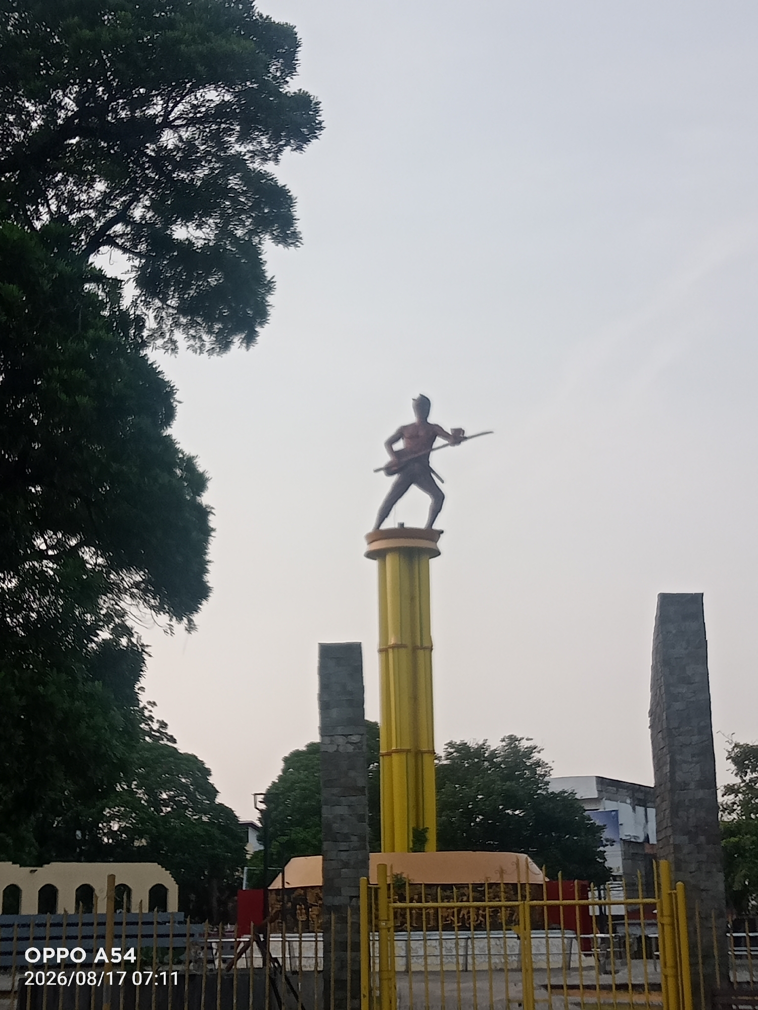The image shows a statue of a warrior holding a spear and shield, standing atop a tall yellow pillar in the city of Jambi, Indonesia. The statue is silhouetted against an overcast sky. To the left of the statue, a large tree with dark green foliage obscures a portion of the sky. To the right of the statue, two tall, grey stone pillars stand, one on each side. In the foreground, a yellow metal fence encloses the area, with some buildings and more trees visible behind it. The timestamp "OPPO A54 2026/08/17 07:11" is visible in the lower left corner of the image.