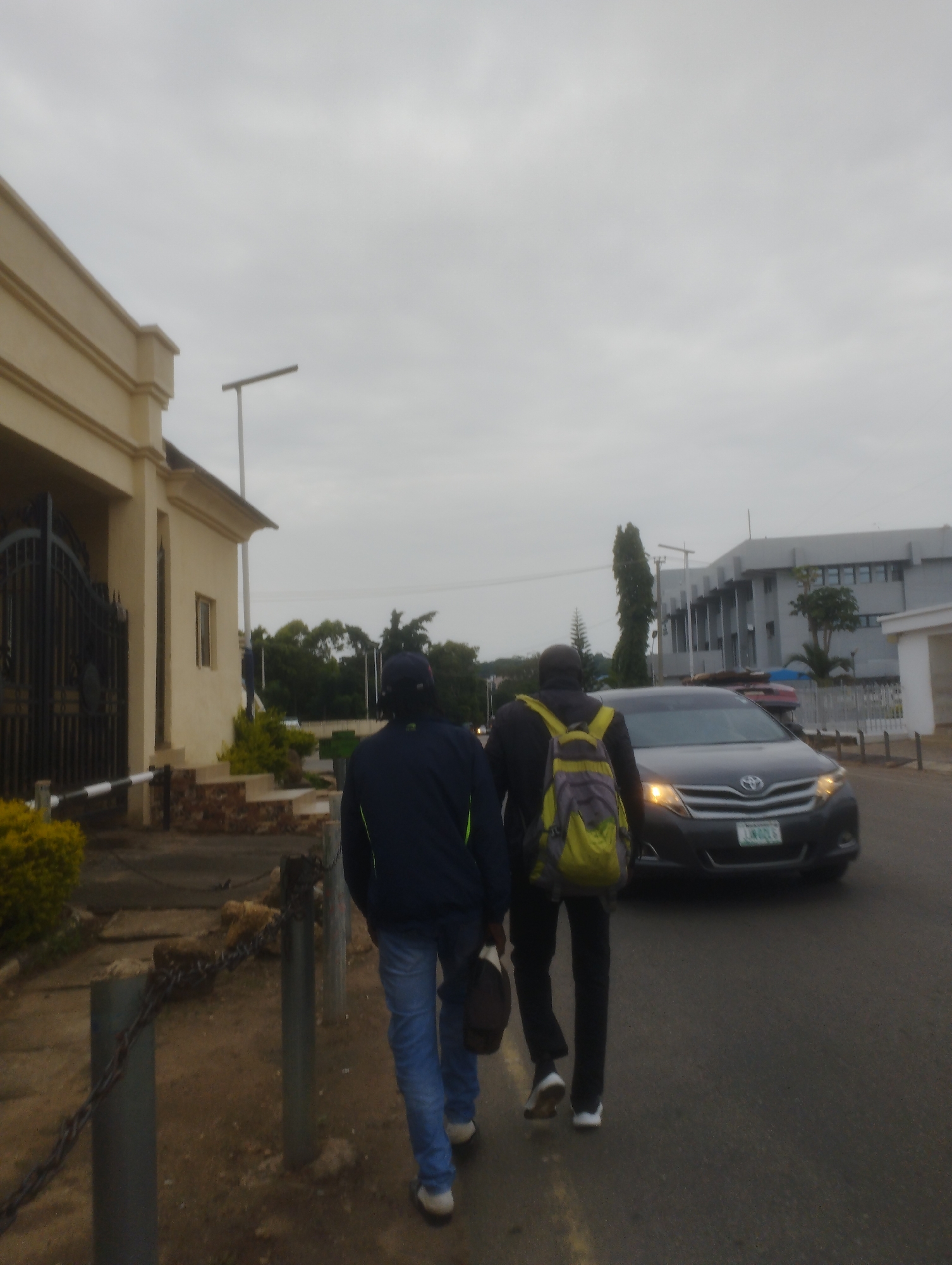 Two individuals walk away from the camera on a paved road in Jos, Nigeria, under an overcast sky. The person on the left wears a dark jacket and blue jeans, carrying a dark bag. The person on the right, with a lime green and grey backpack, wears a dark hooded jacket and dark pants. To the left of the road is a cream-colored building with a large black gate. Further down the road, a dark gray Toyota minivan drives towards the camera. In the background, several modern buildings and trees are visible. The ground beside the road is dirt and gravel, with a metal chain fence lining the area.