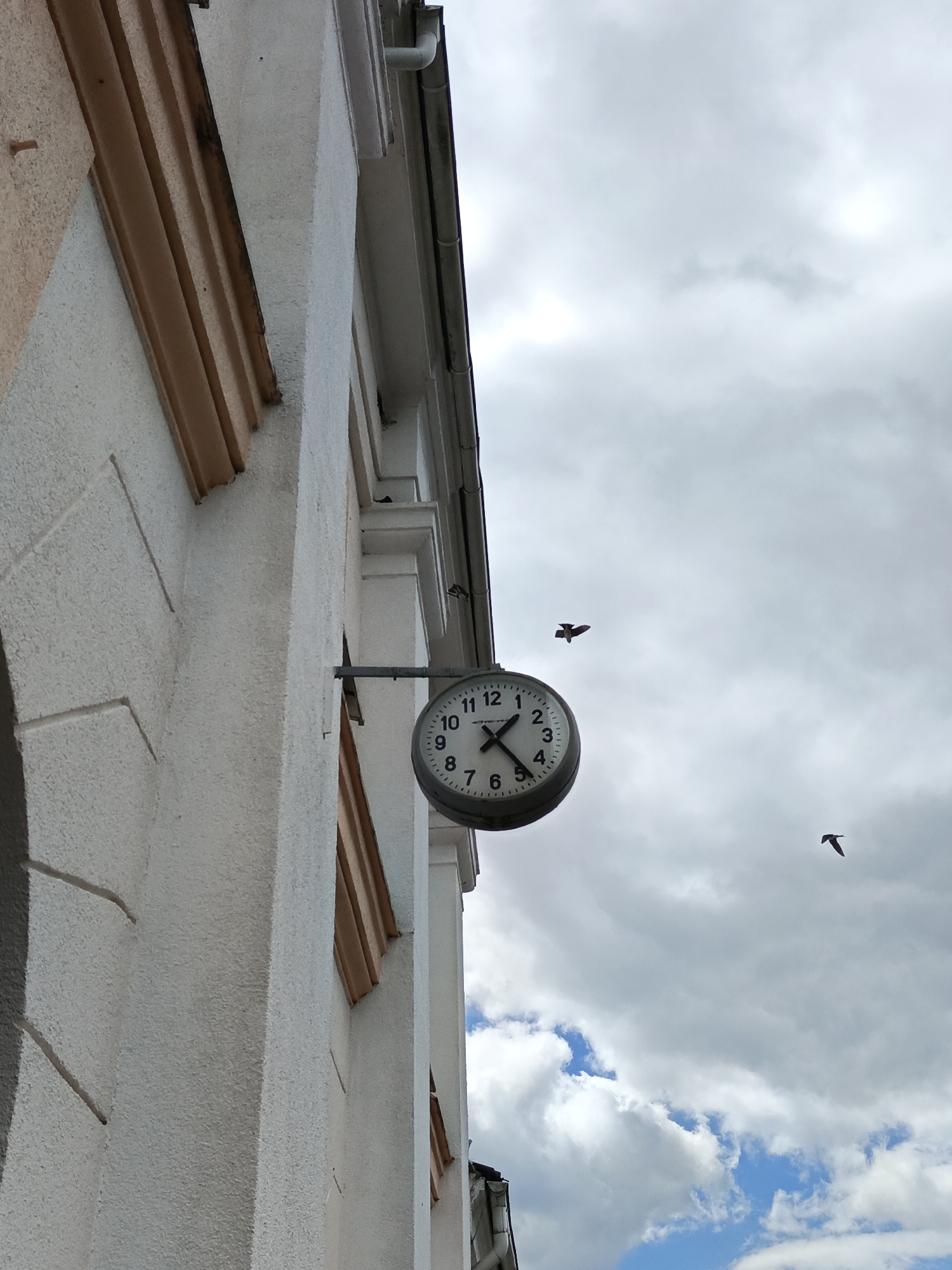A round analog clock hangs on the exterior wall of a building. The clock face is white with black numbers and hands. The time appears to be approximately 10:10. Two birds are captured in flight in the sky, which is partly cloudy. The building appears to be made of white stucco with decorative beige trim. The scene is likely outdoors, possibly in a town or city setting in Belarus, given the location context. The weather appears overcast with some patches of blue sky and sunlight filtering through the clouds. No visible text is present on the clock or building.