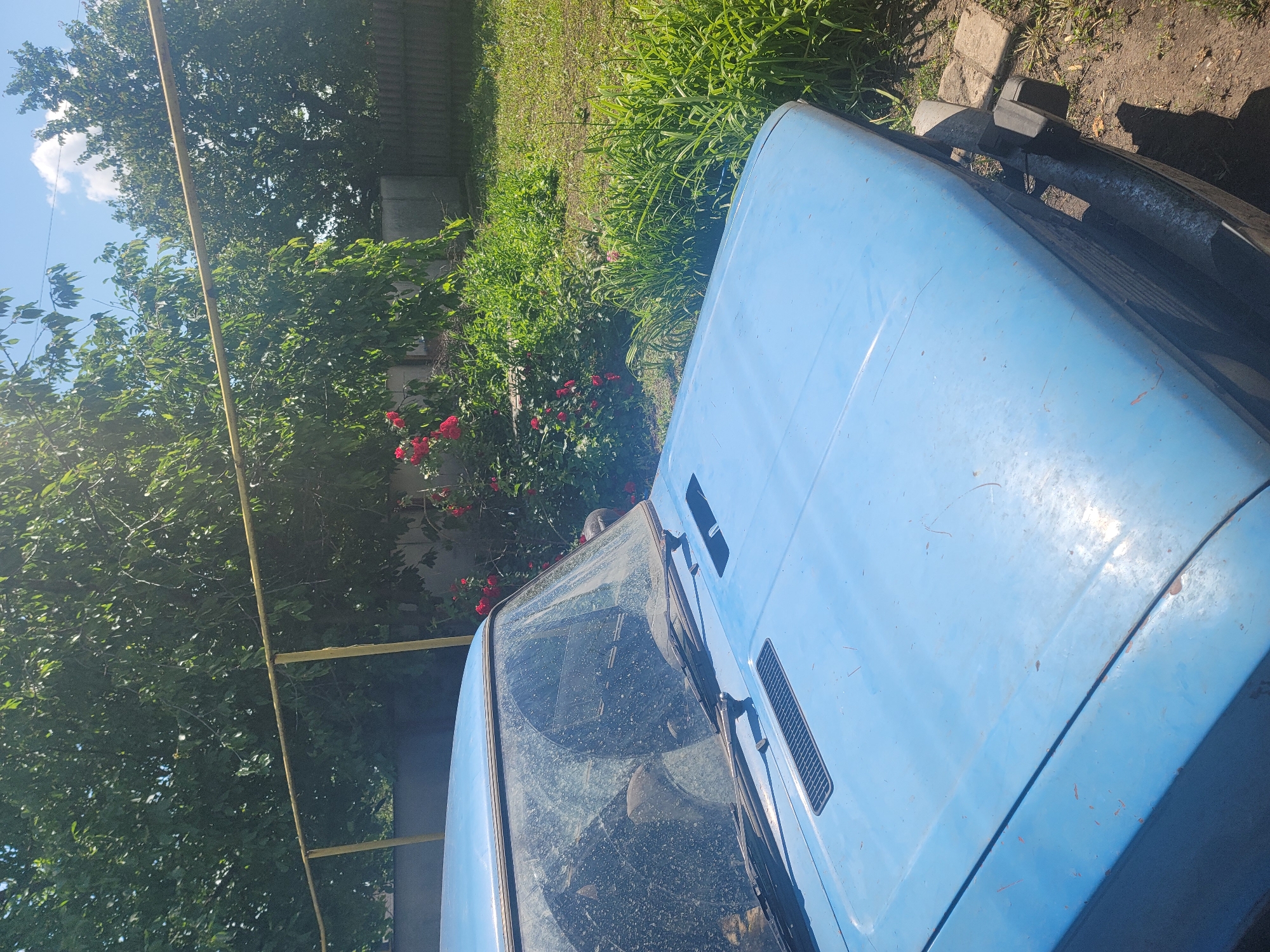 The image shows the front of a light blue, older model car parked on a dirt surface with some scattered stones and grass. The car's hood is clean, but the windshield is heavily covered in dust and dirt. Behind the car, a lush green garden area with bright red roses in bloom is visible. A yellow metal pipe runs horizontally across the upper portion of the image, supported by posts. In the background, trees and a wooden fence can be seen, suggesting a rural or suburban setting. The sky is blue with some white clouds. The overall impression is that of a car parked in a yard on a sunny day in Vovkivka, Ukraine.