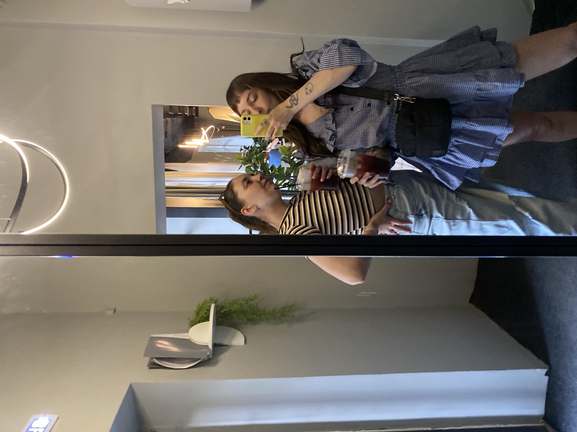 Two women are reflected in a mirror. One woman is wearing a blue and white checkered dress with a black purse, and she is holding a yellow smartphone to take a picture. The other woman is wearing a striped shirt and denim jeans, and she is holding two drinks. The background features a light fixture, plants, and a grey wall. The location in the city of Votkinsk, Russia is not identifiable from the image.