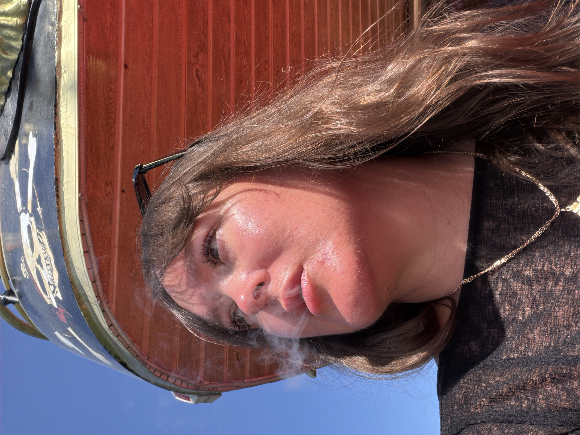 A young woman is smoking, exhaling smoke from her lips. She is wearing glasses and a gold necklace. The background shows a blue sky and a carnival ride with a skull and crossbones design. The location is likely Alanya, Turkey, given the style of the carnival ride and the lighting.