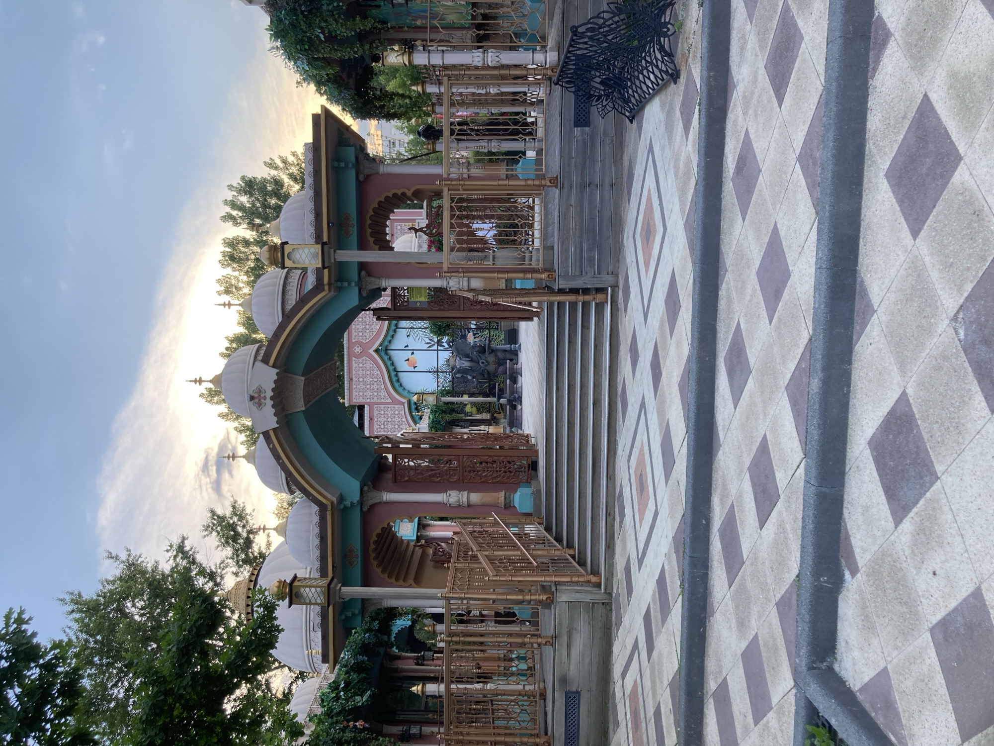 An outdoor shot captures a decorative entrance structure with multiple domed roofs, reminiscent of Indian architecture. The structure is painted in a combination of pink, teal, and gold, with intricate metalwork on the railings and gates. A wide, checkered tiled walkway leads up to a set of stairs that access the entrance. In the background, trees and a cloudy sky are visible, suggesting it is late afternoon or early evening. The location is identified as Moscow, Russia, specifically within the Ostankino district.
