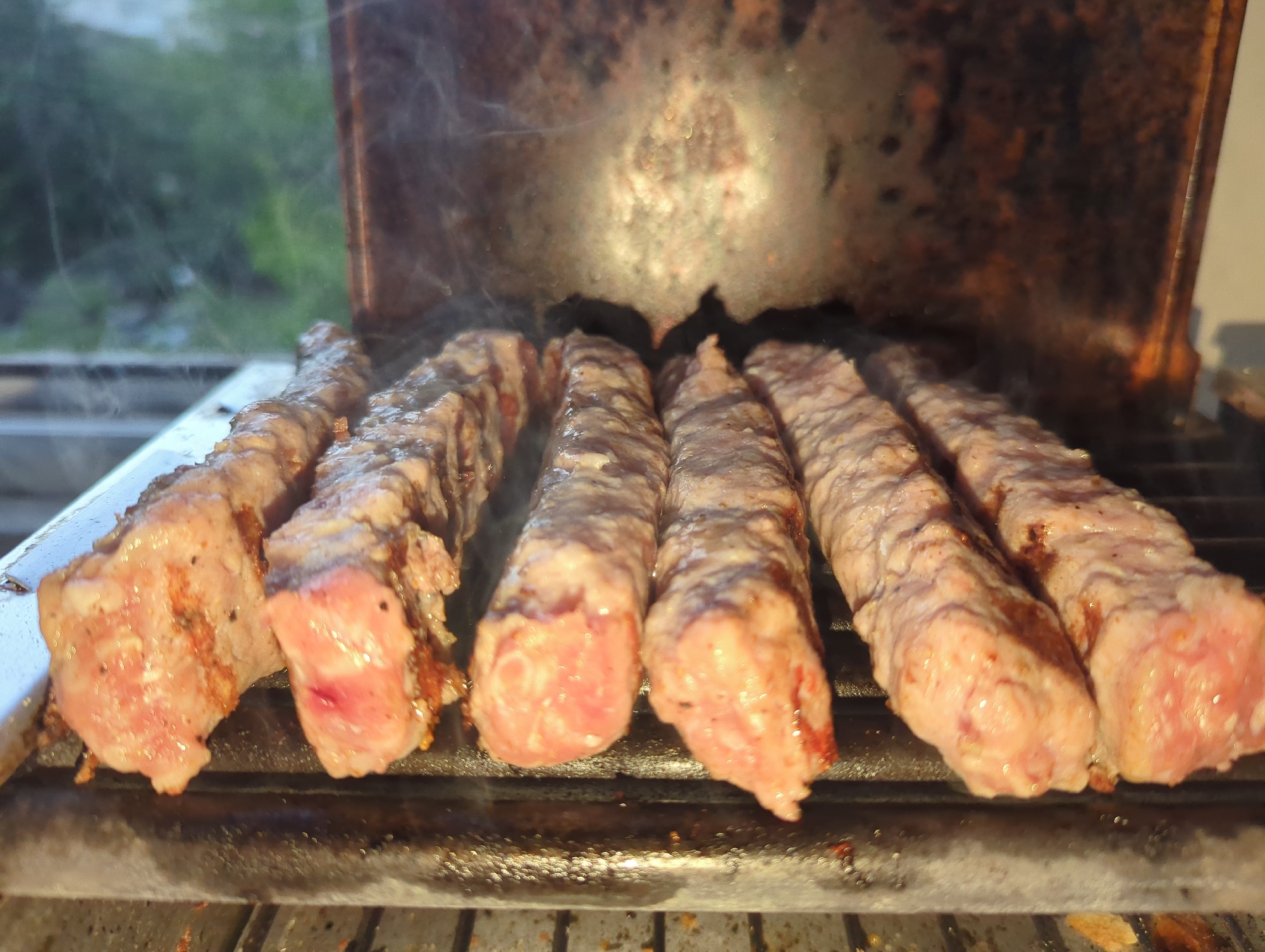 The image shows several cevapcici, a grilled dish of minced meat, cooking on a grill. The meat is lightly browned and still appears pinkish in some areas, suggesting it is not fully cooked. Smoke rises from the grill, indicating that the food is actively being cooked. The grill itself appears to be a smaller, portable model, possibly used outdoors. The background is blurred but suggests an outdoor environment, potentially a backyard or patio, with foliage visible in the distance. The image does not contain enough information to place it definitively in Varna, Bulgaria. There are no people visible in the image.