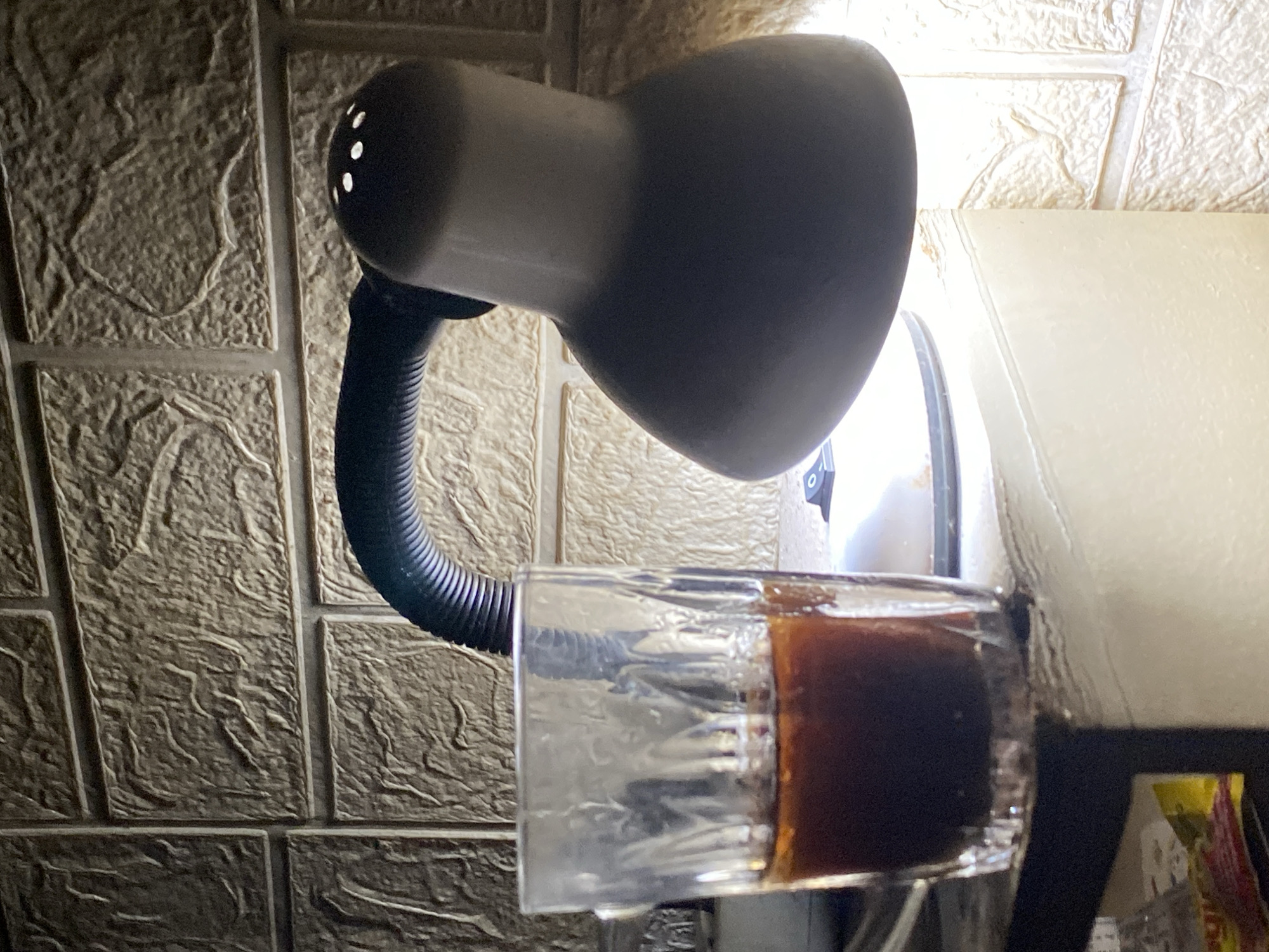 An image shows a glass of coffee next to a desk lamp, placed on a white surface, with a brick-patterned wall in the background. The scene appears to be indoors, likely a home or office setting, given the presence of a desk lamp and coffee. The lighting is dim, with the lamp providing the primary light source, creating shadows and highlighting the texture of the wall. The specific location in Batam, Indonesia, cannot be determined from the visual information alone.