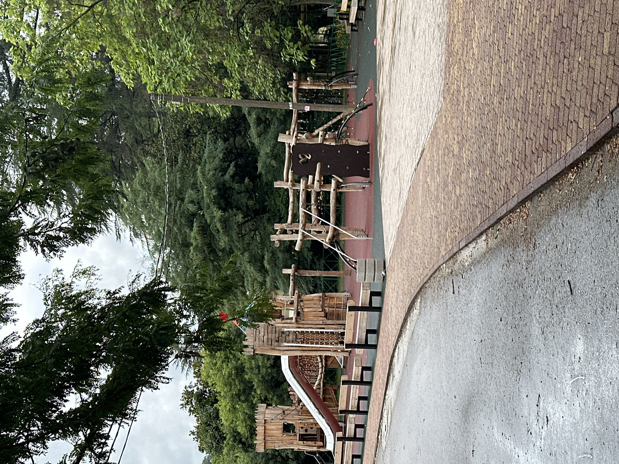 The image depicts a wooden playground in a park, likely located in Sochi, Russia, given the dense, lush greenery and overcast sky characteristic of the region. The playground features a large, castle-like wooden structure with a slide and climbing elements. Benches are arranged in front of the structure, and a paved pathway with a brick pattern curves around the area, bordering a wet, asphalted surface. In the background, a dense forest of tall trees contributes to the park's natural setting. The scene is devoid of people, creating a sense of quiet and stillness. The lighting suggests it is daytime, but the cloudy conditions cast a diffused, even light over the entire scene.
