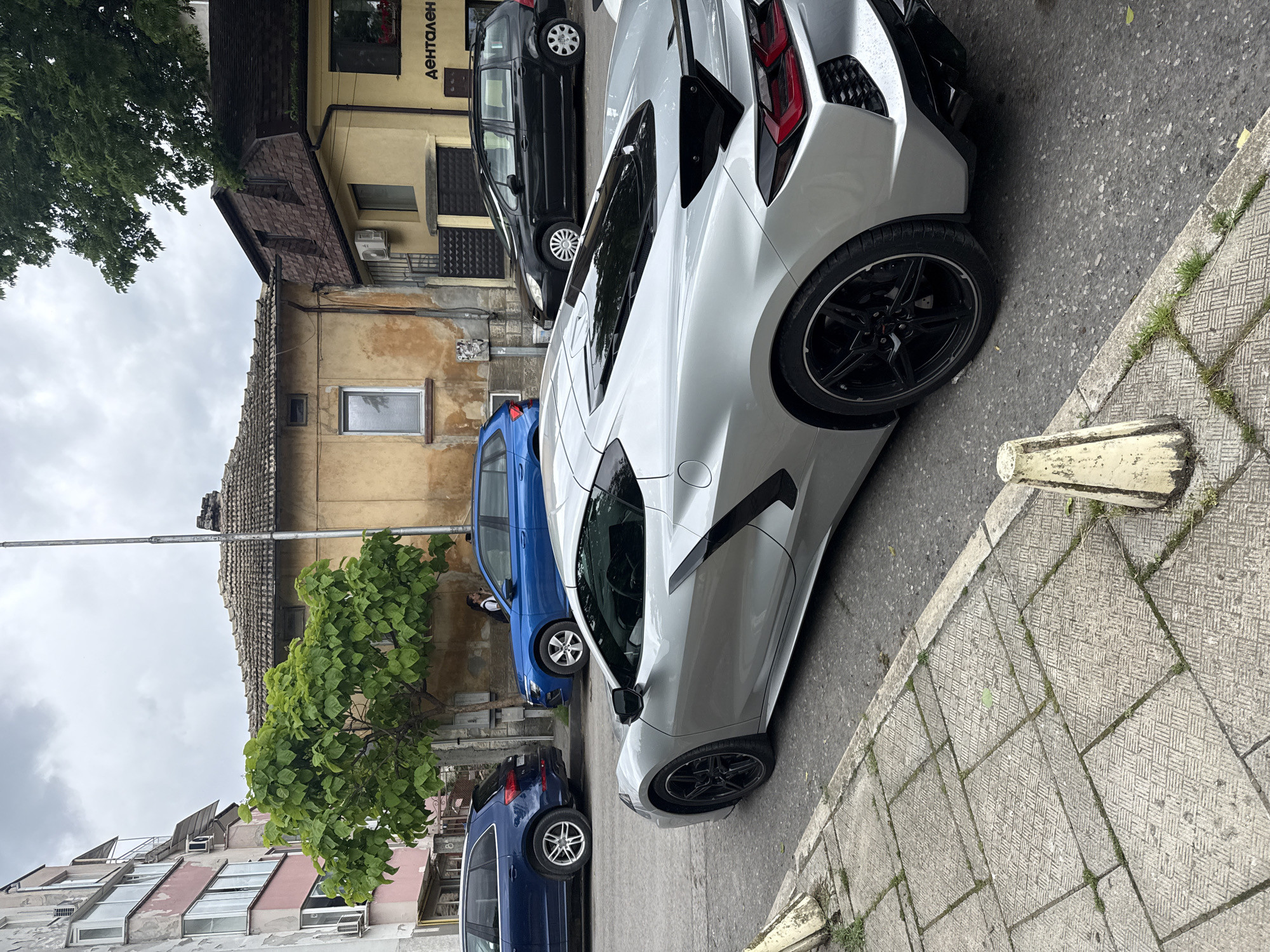 This daytime image captures a street scene in Shumen, Bulgaria, under an overcast sky. The photo is rotated 90 degrees counter-clockwise.
In the foreground and mid-ground, a striking light silver/grey Chevrolet Corvette C8 sports car is prominently parked on the right side of the street, facing away from the viewer. Its modern design, distinctive rear lights, and black multi-spoke alloy wheels are clearly visible. Behind it, a blue hatchback is parked, followed by another dark blue SUV/wagon. On the opposite side of the street, a black minivan-style car, possibly a Peugeot or Citroën, is parked alongside the curb.
The street is paved with asphalt, and a patterned concrete sidewalk, showing some wear and interspersed with small patches of grass, runs along the right. A yellow and white painted concrete barrier or post sits at the edge of this sidewalk.
The buildings lining the street present a mix of ages and styles. On the left side (when upright), an older building with a faded yellow/beige facade features the visible Cyrillic text "ДЕНТАЛЕН" (meaning "DENTAL" in Bulgarian), indicating a dental establishment. Its roof is covered with brown tiles. Further down the street, other residential and commercial buildings are visible, some with multiple windows and air conditioning units, and tiled or flat roofs. A large tree with abundant green foliage dominates the upper left part of the scene, adding natural elements to the urban environment. The overall impression is that of a quiet, everyday street in a Bulgarian town.