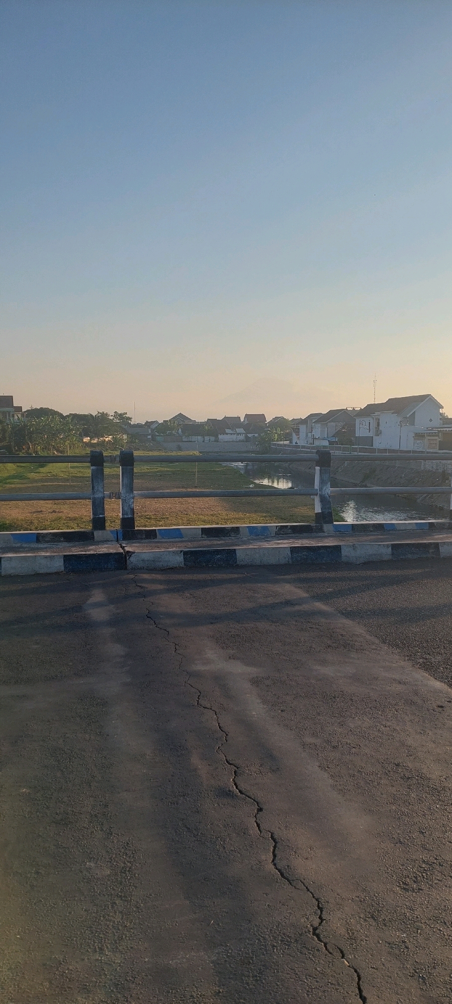 The image depicts a serene scene of a residential area with a river or canal running through it, viewed from a bridge in Lumajang, Indonesia. The sky is a clear, pale blue, suggesting it is either early morning or late afternoon. In the foreground, a cracked asphalt road occupies the lower portion of the frame.
The bridge railing, made of dark grey posts with a metal bar, runs horizontally across the mid-section of the image. Below the railing, there is a concrete curb painted with alternating black and blue stripes. A narrow body of water, likely a canal or small river, is visible beyond the bridge, reflecting the soft light.
On the other side of the water, a green grassy area leads up to a row of houses. These houses have tiled roofs and appear to be of various sizes and designs, typical of a suburban neighborhood. Some houses have visible windows and walls, and vegetation, including trees and bushes, is interspersed throughout the area. In the distance, a faint outline of a mountain can be seen against the horizon, partially obscured by haze. The overall impression is one of a quiet, rural or semi-rural setting within Lumajang. There are no people visible in the image.