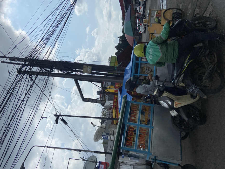 The image captures a street scene in Padalarang, Indonesia (latitude: -6.856, longitude: 107.498). A man wearing a green jacket with "Grab" emblazoned on the back is seated on a black motorcycle, looking right, holding the handlebars. The motorcycle is parked near a blue and white food cart, with a sign in Indonesian that reads "BASO TAHU SIDMAY" (Meatball, Tofu, and a type of Indonesian snack). Another man, wearing a gray long-sleeve shirt, is standing near the food cart, looking toward the motorcycle. Behind the motorcycle, a white food cart is being unloaded. The background is a jumble of power lines, a few buildings, and trees. The ground is paved with asphalt. The day is overcast and the sky is grey with a few white clouds.