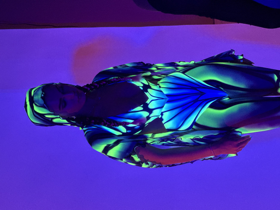 A female individual is centrally depicted in an interior setting illuminated by ultraviolet light, located in Sofia, Bulgaria. She stands facing slightly right, with her head bowed and gaze directed downwards. Her hair is styled in two braids, appearing dark with faint pinkish fluorescence. She wears a multi-piece costume designed to fluoresce, featuring prominent neon green and blue patterns resembling butterfly wings. The attire includes a plunging V-neck top or bodysuit, matching lower body coverage, a long-sleeved open-front outer garment with ruffled cuffs, and a matching head covering. A delicate necklace is visible on her neck, and a thin bracelet adorns her left wrist. The background is a solid, deep violet-blue, with a vertical light reddish-purple strip visible on the far right, indicating a structural element such as a corner or panel. An indistinct, dark form is partially visible on the extreme lower right. The scene's primary illumination source causes the garment's specific dyes to emit light.
